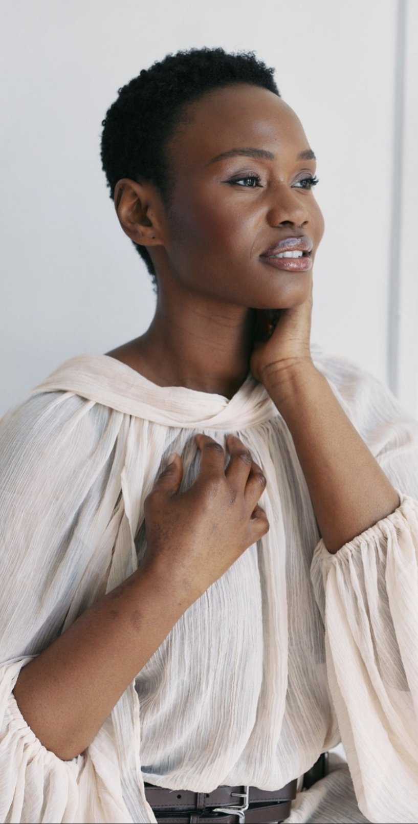 A woman with short curly hair looking contemplative, wearing a cream-colored blouse, touching her neck and chest, standing against a plain background.
