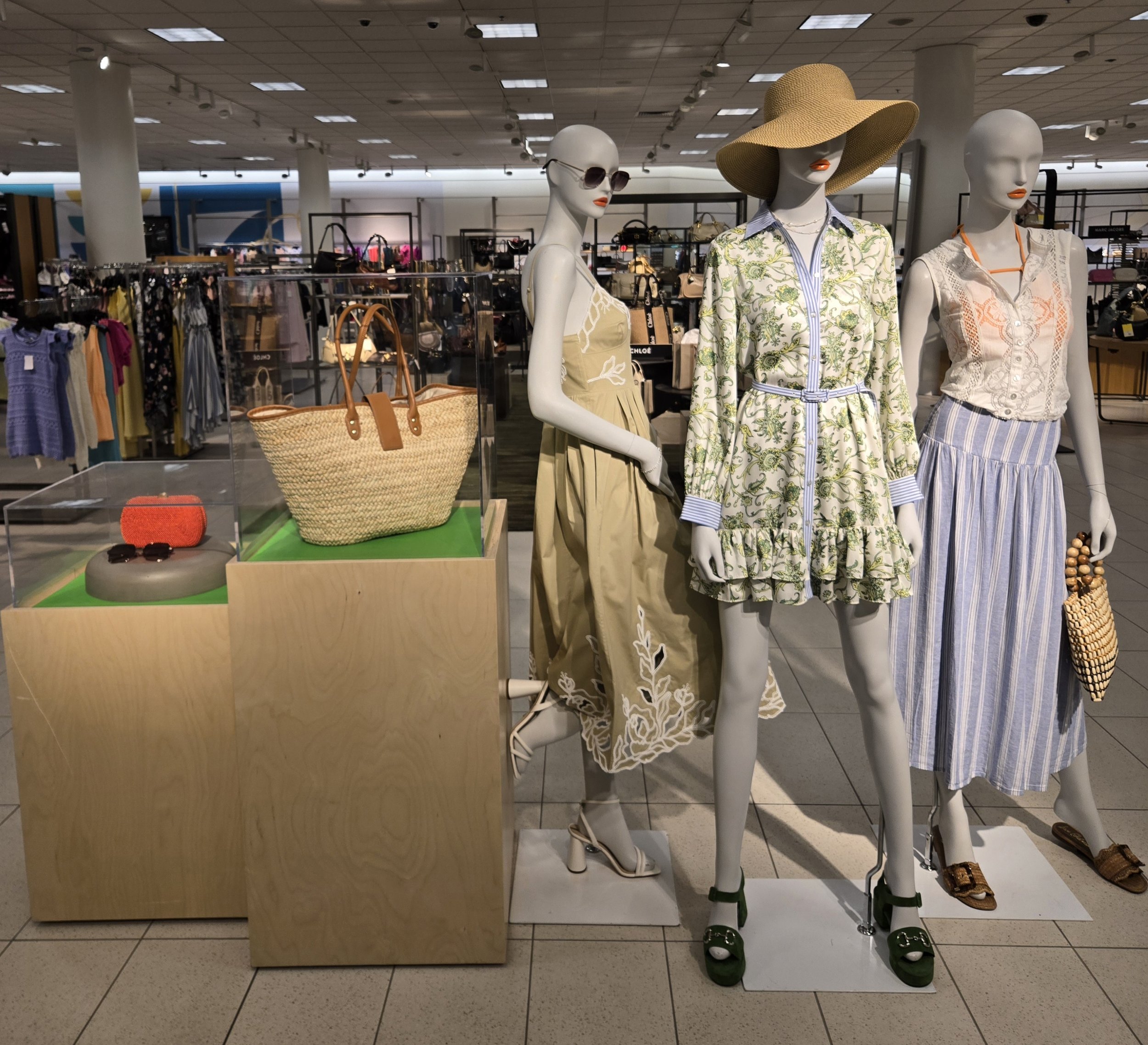 Display of three mannequins dressed in summer clothing in a retail store. Mannequin on the left wears sunglasses and a beige dress with floral details. Mannequin in the middle wears a wide-brimmed sun hat and a light-colored dress with a botanical print. Mannequin on the right is dressed in a sleeveless top and a striped skirt, also with a wide-brimmed hat. The background features shelves with handbags and clothing racks.