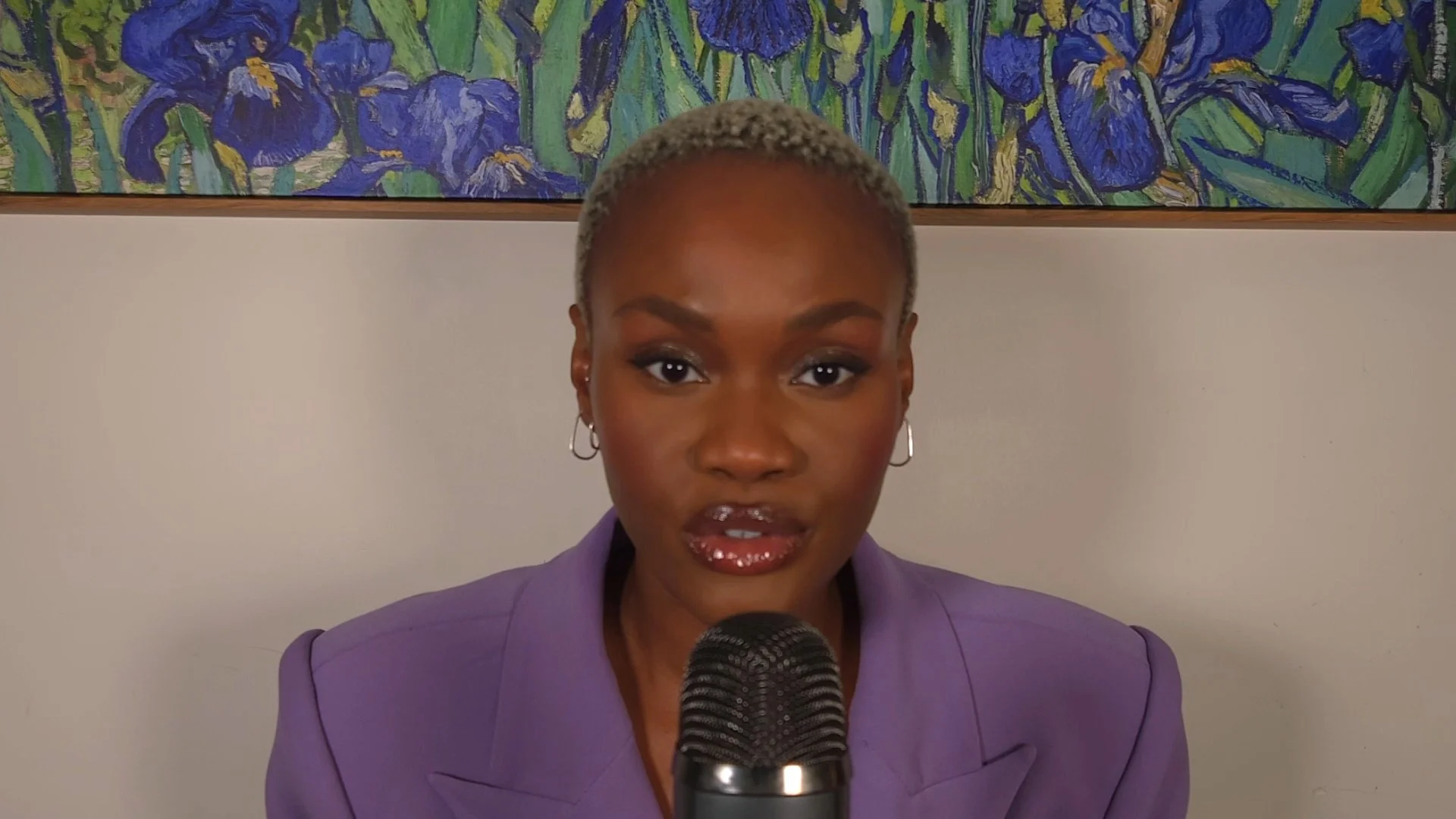 Hilary Biaku | Black Female Model, Creative Strategist, Content Creator, Public Speaker, A woman with short, light-colored hair, wearing a purple blazer and hoop earrings, speaking into a microphone with a colorful, floral painting in the background.