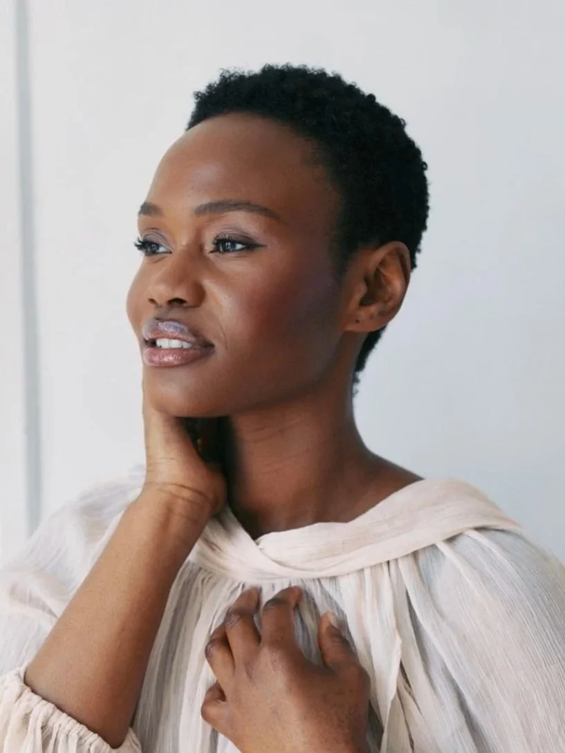 Hilary Biaku | Creative Strategist, Model, Content Creator - A portrait of a woman with short black curly hair, wearing a light-colored, pleated blouse, gently touching her neck and chest with a thoughtful expression, against a plain background.