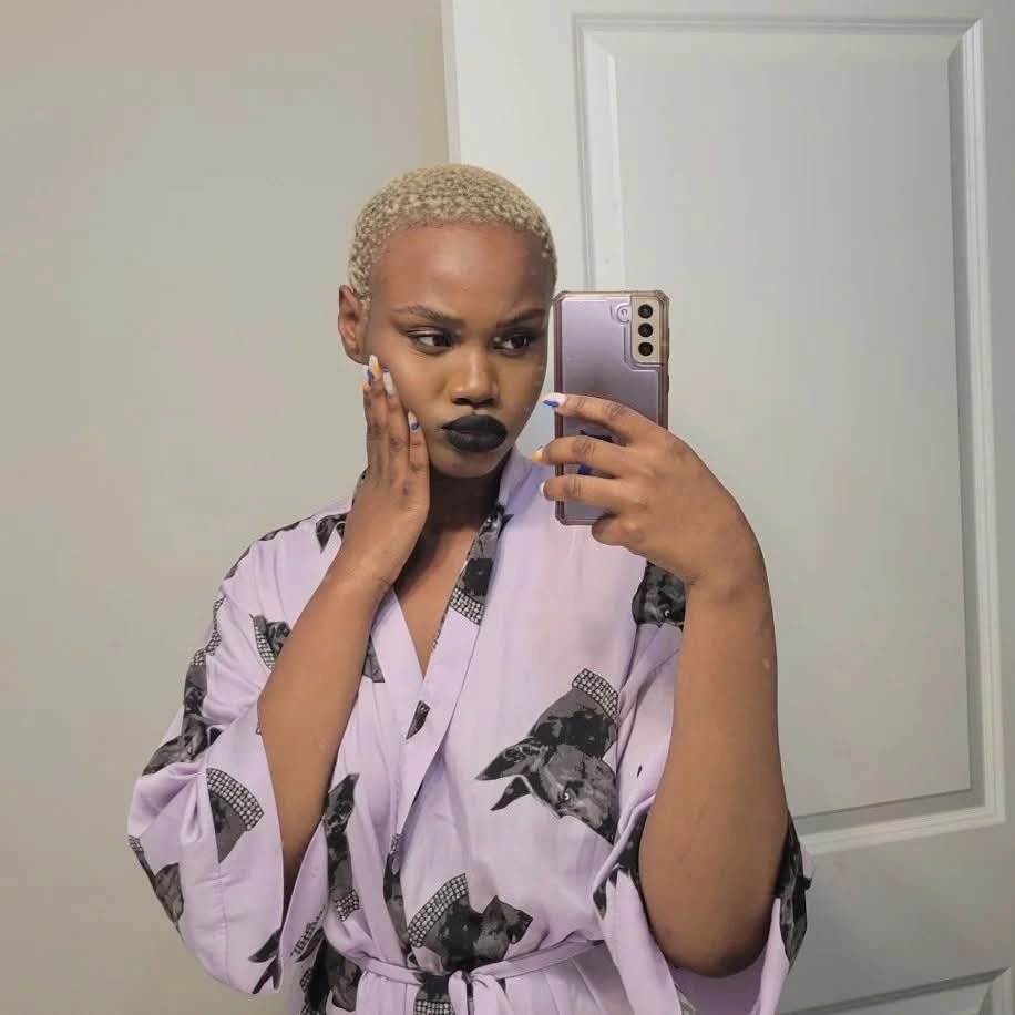 A woman with short blonde hair taking a mirror selfie indoors, wearing a pink robe with black cat patterns, with a serious expression and dark lipstick.