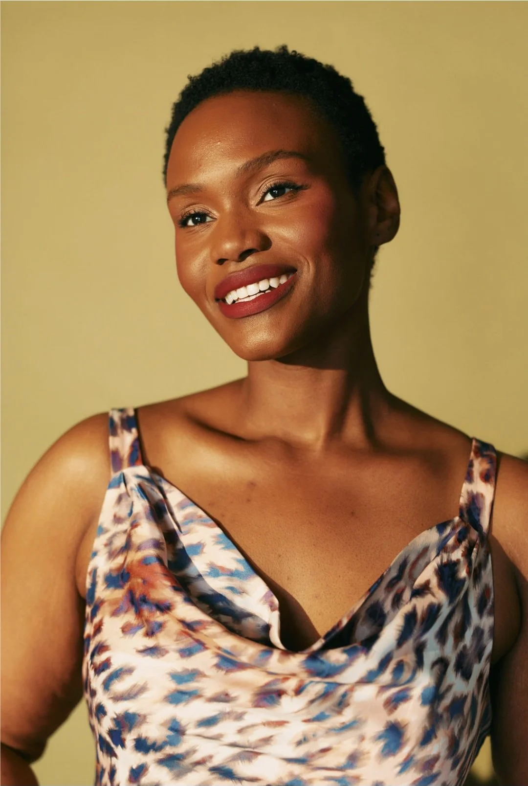 Hilary Biaku | Creative Strategist, Model, Content Creator, Public Speaker - A Black woman with short, curly black hair smiling, wearing a sleeveless, multicolored dress with a cowl neckline, standing against a plain, yellow-green background.