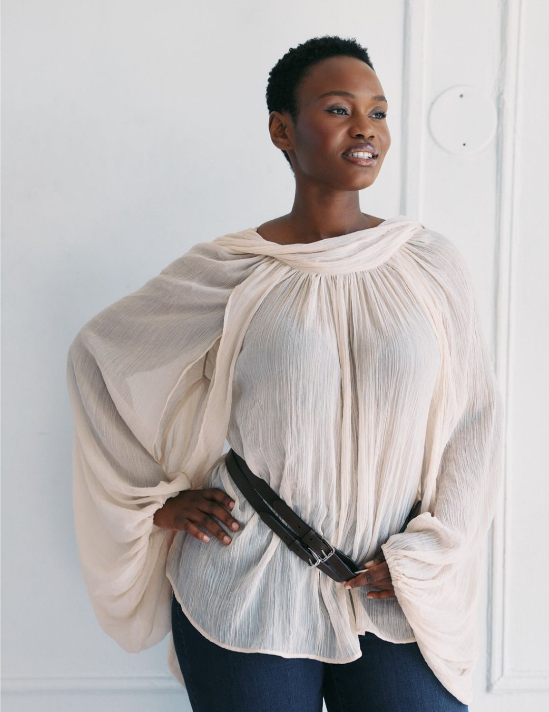 Hilary Biaku | Black Female Model, Creative Strategist, Content - A woman with short curly hair, wearing a loose, cream-colored pleated blouse, dark jeans, and a black belt, standing with one hand on her hip and looking to her left, smiling softly.