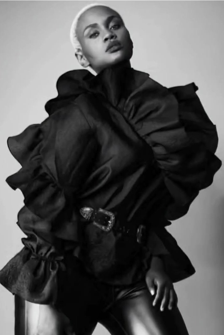 Hilary Biaku | Black Female Model, Creative Strategist, Content Creator, Public Speaker, and Black and white photo of a woman wearing a voluminous high-fashion jacket with oversized sleeves and a fur-trimmed belt, standing against a plain background.