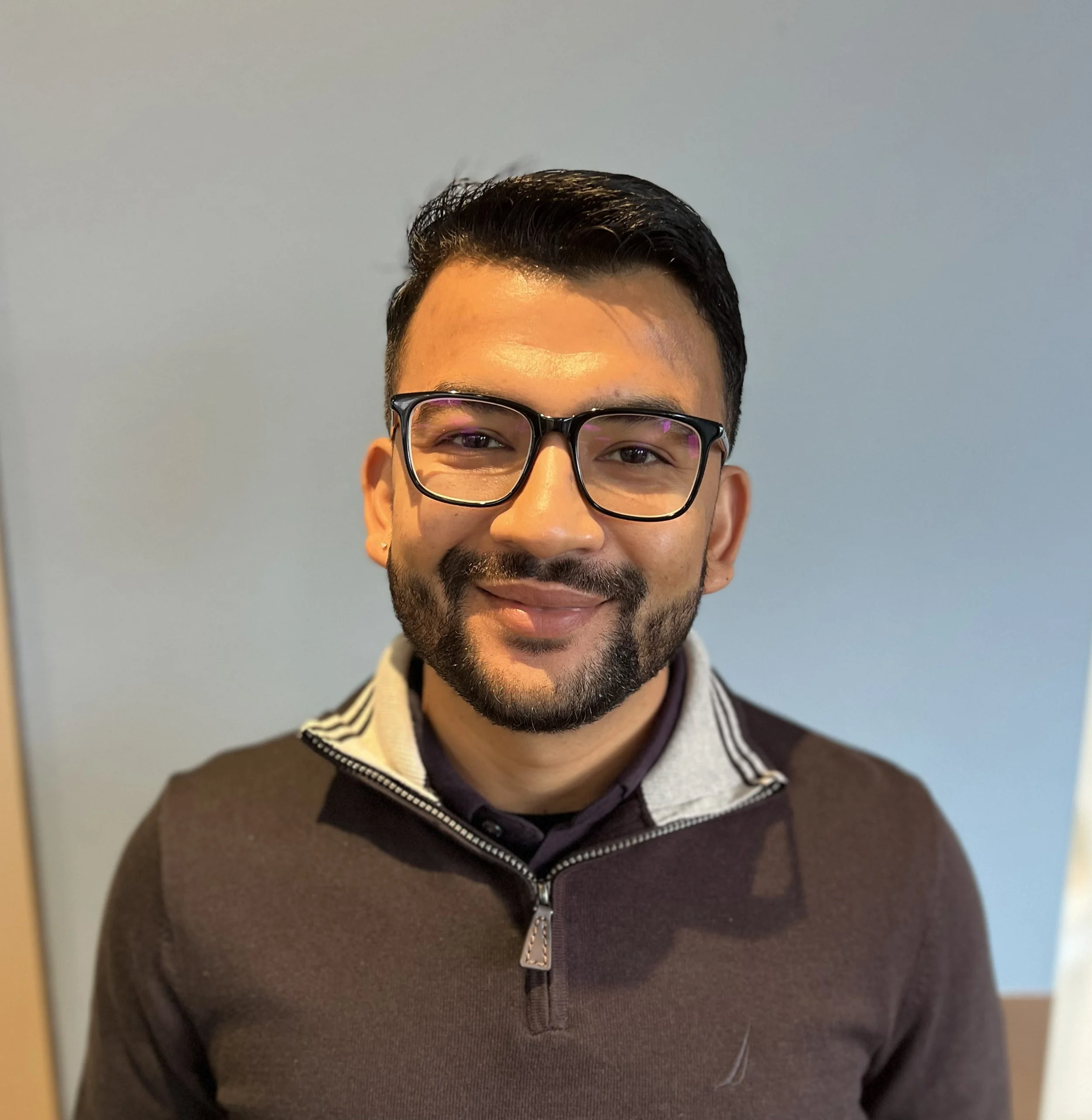 Close-up portrait of a smiling man with glasses, dark hair, and a beard, wearing a dark pullover with a half-zip, standing against a blurred indoor background.