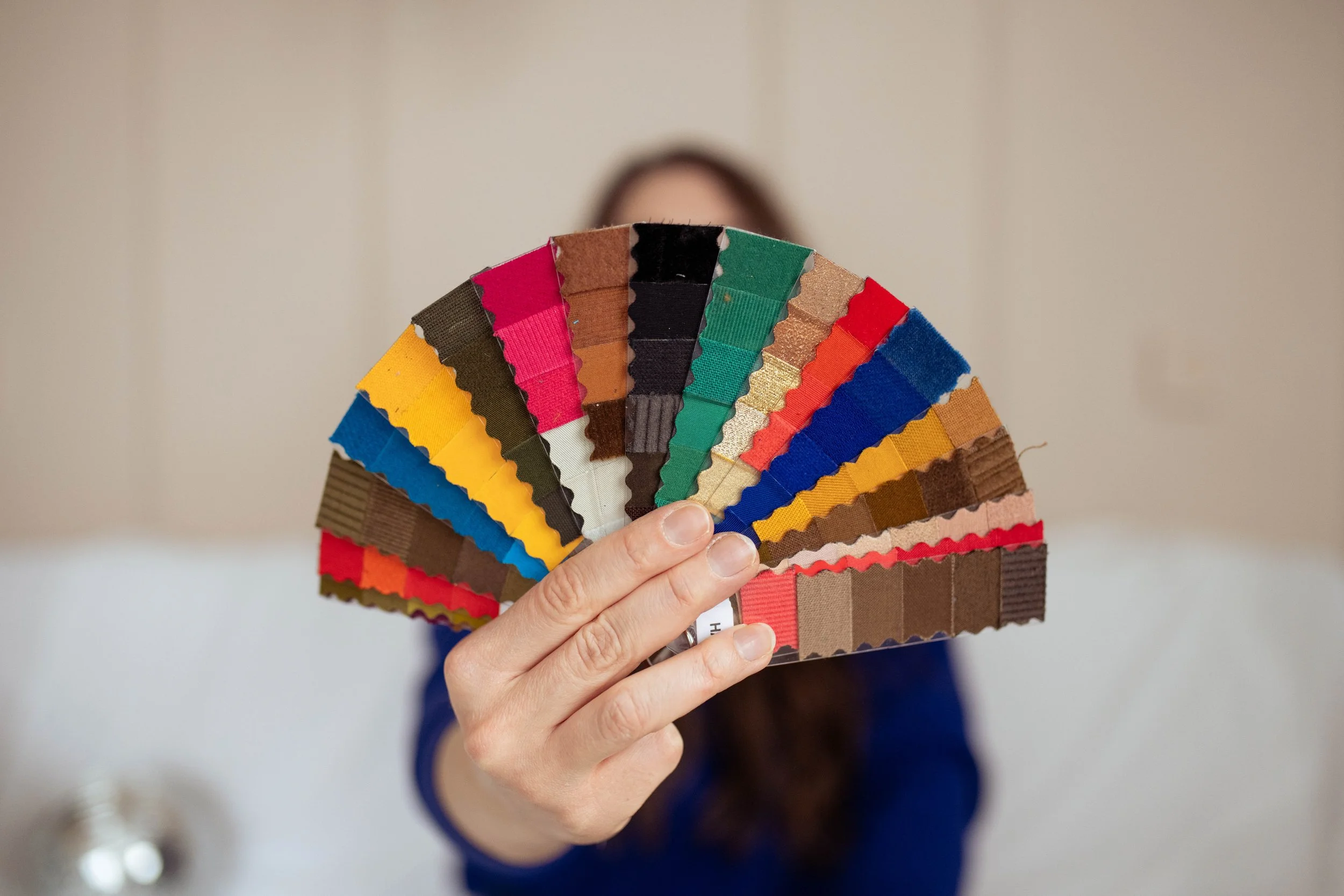 Person holding a fan of a custom color analysis fan made of fabric swatches in various colors and textures, partially obscuring their face.