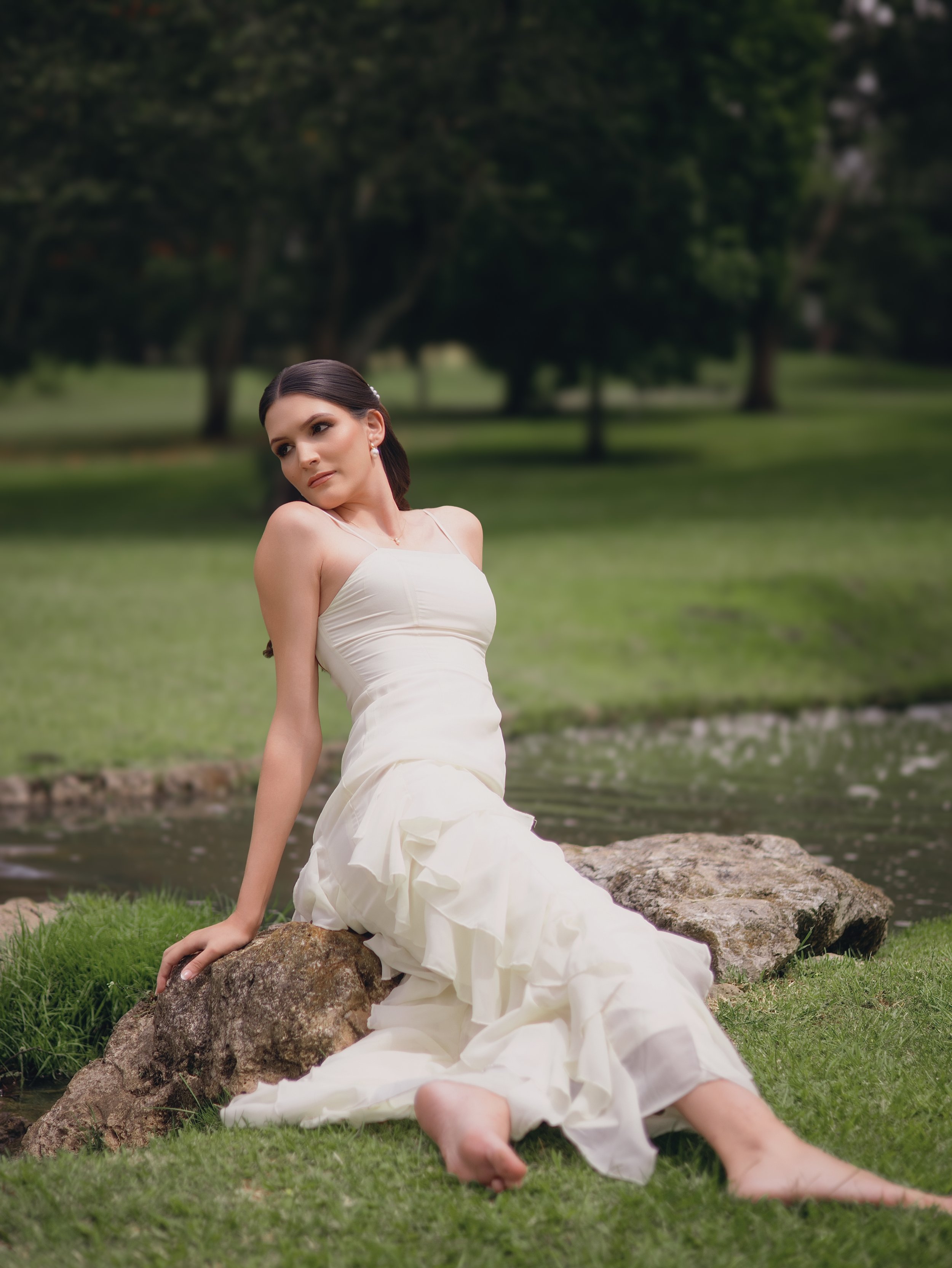 Mujer con vestido blanco en un entorno natural con árboles y césped, sentada sobre rocas junto a un río o lago