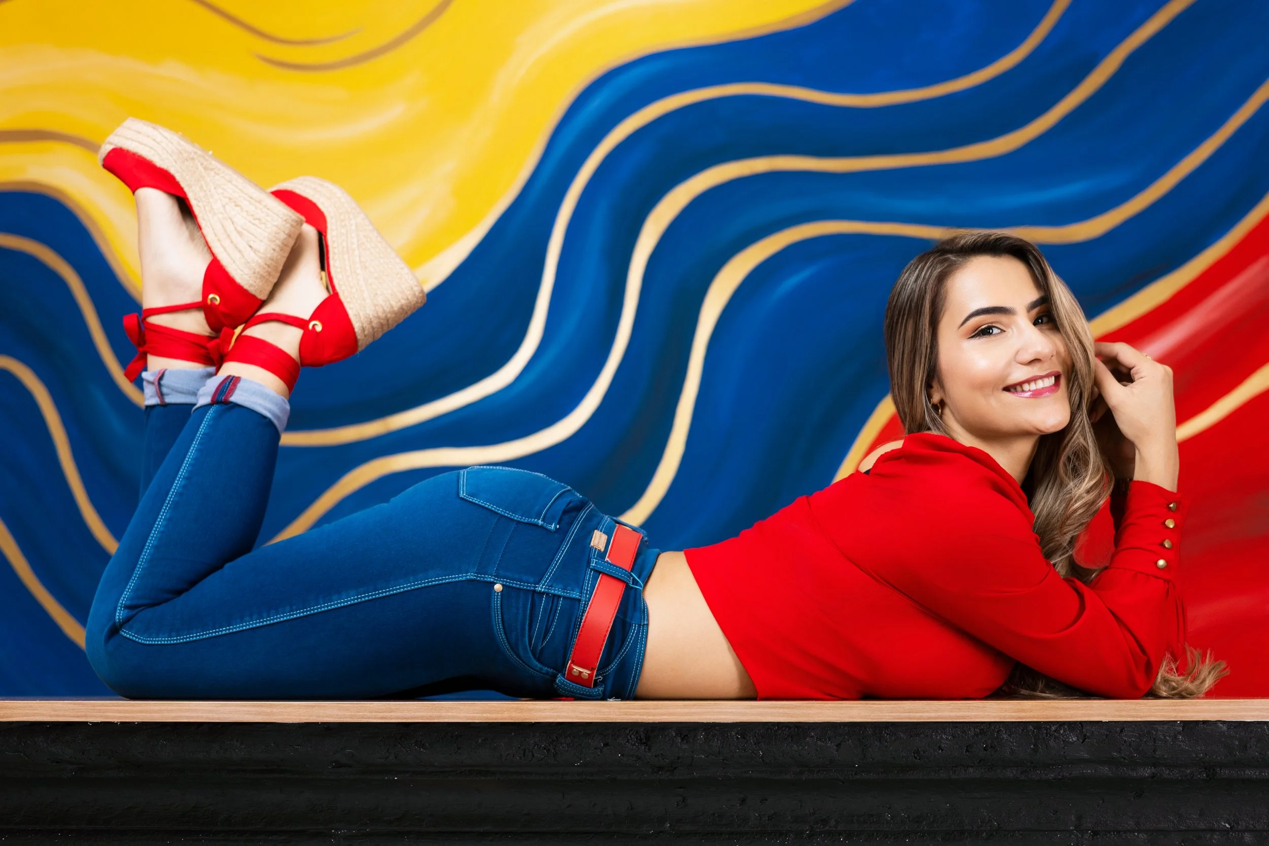 Joven mujer acostada en una mesa, vestida con pantalones azules, suéter rojo y zapatos rojos con plataforma de esparto, frente a un fondo de pared decorativa amarilla, azul y roja.