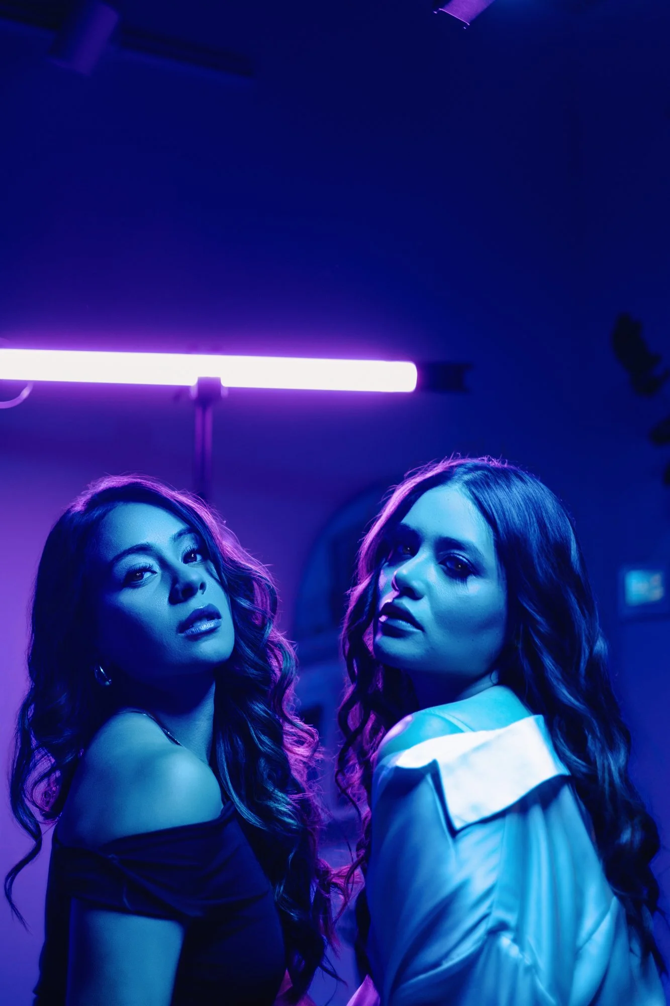 Dos mujeres con cabello rizado y maquillaje oscuro posando bajo una luz de neón púrpura en un ambiente oscuro.