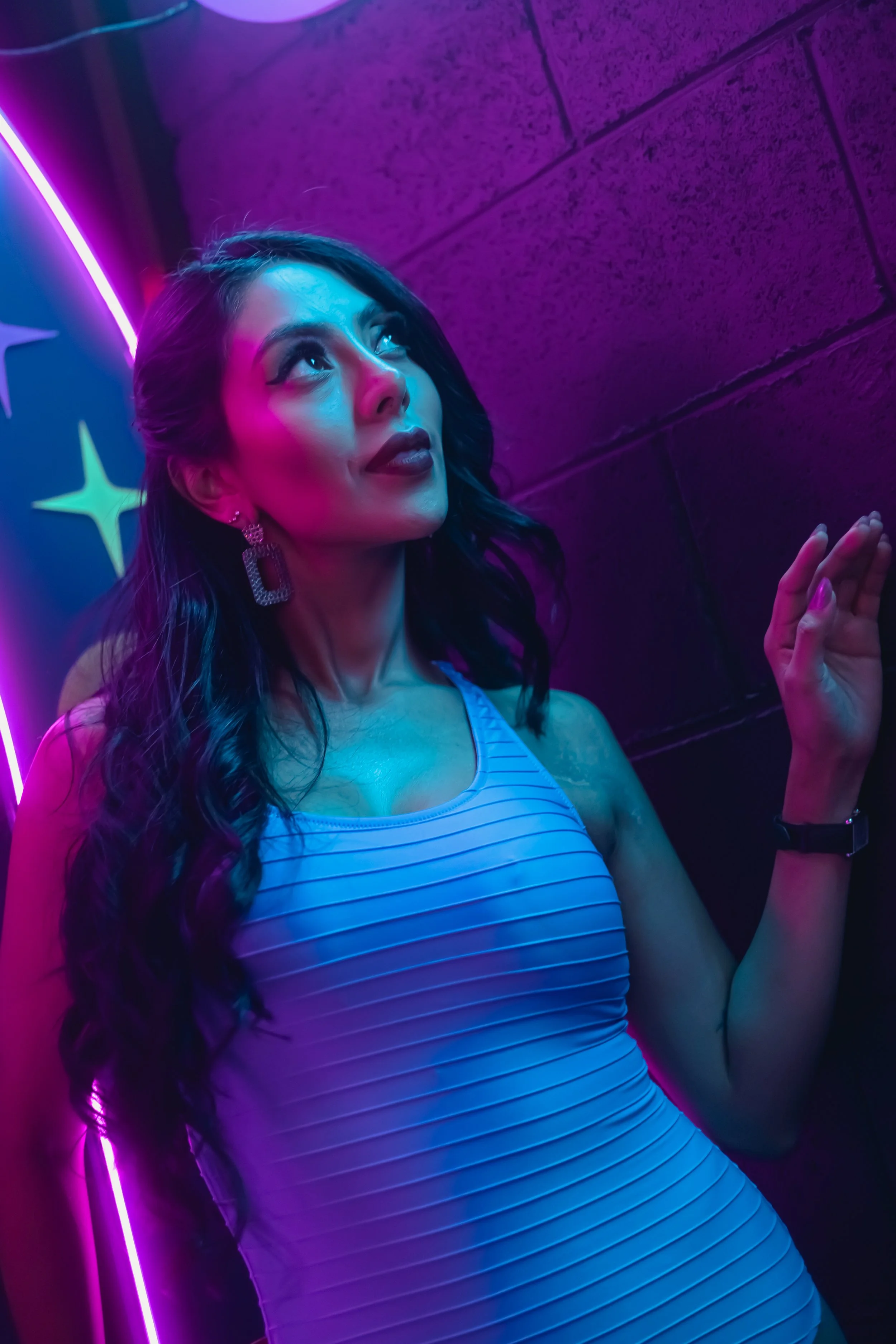Una mujer con cabello oscuro, vestido ajustado de rayas blancas y azules, y pendientes grandes, posa contra una pared con luces de neón rosas y moradas en un entorno con ambiente moderno y vibrante.