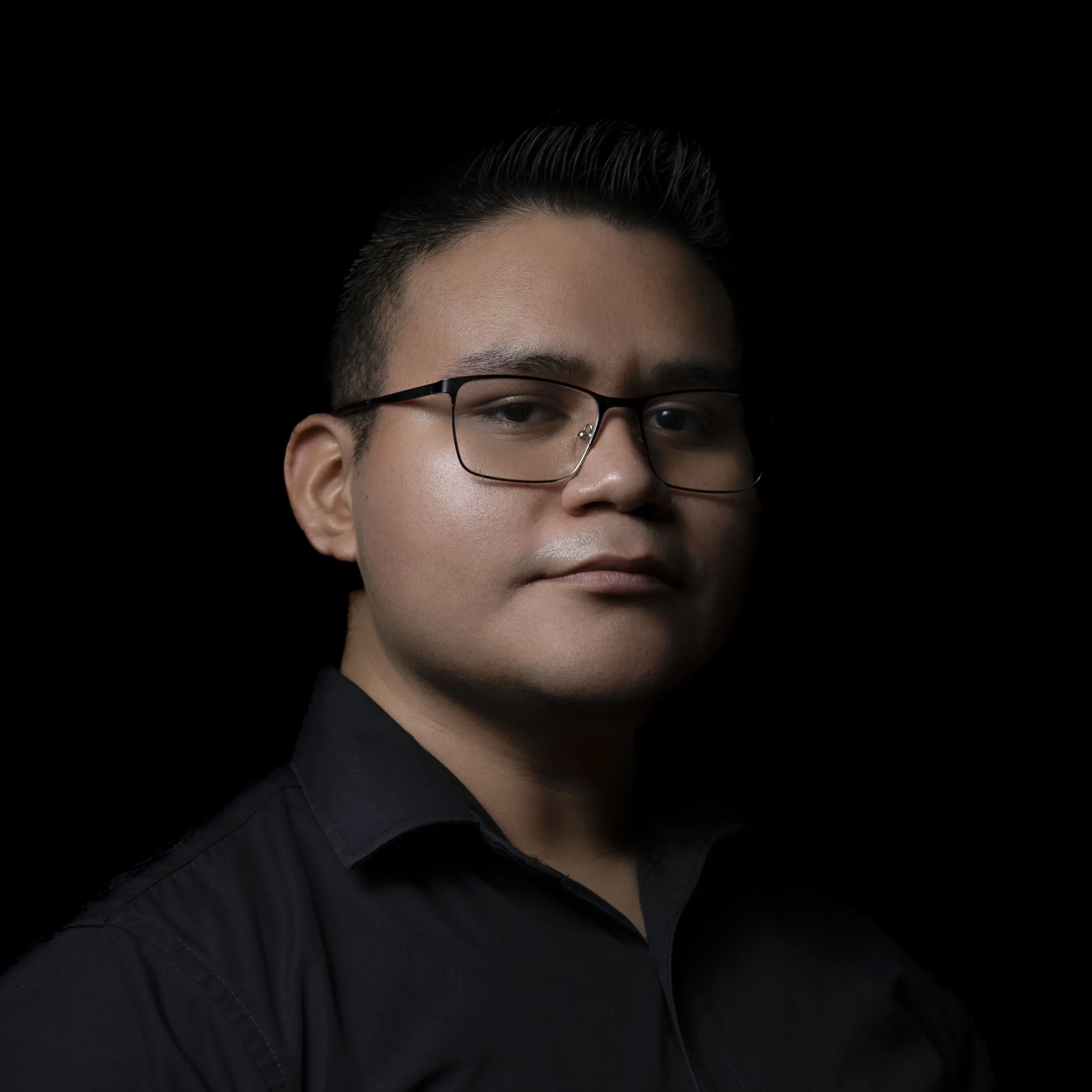 Retrato de un hombre con gafas, cabello bien peinado, piel clara y expresión seria, vestido con camisa negra, en fondo negro.
