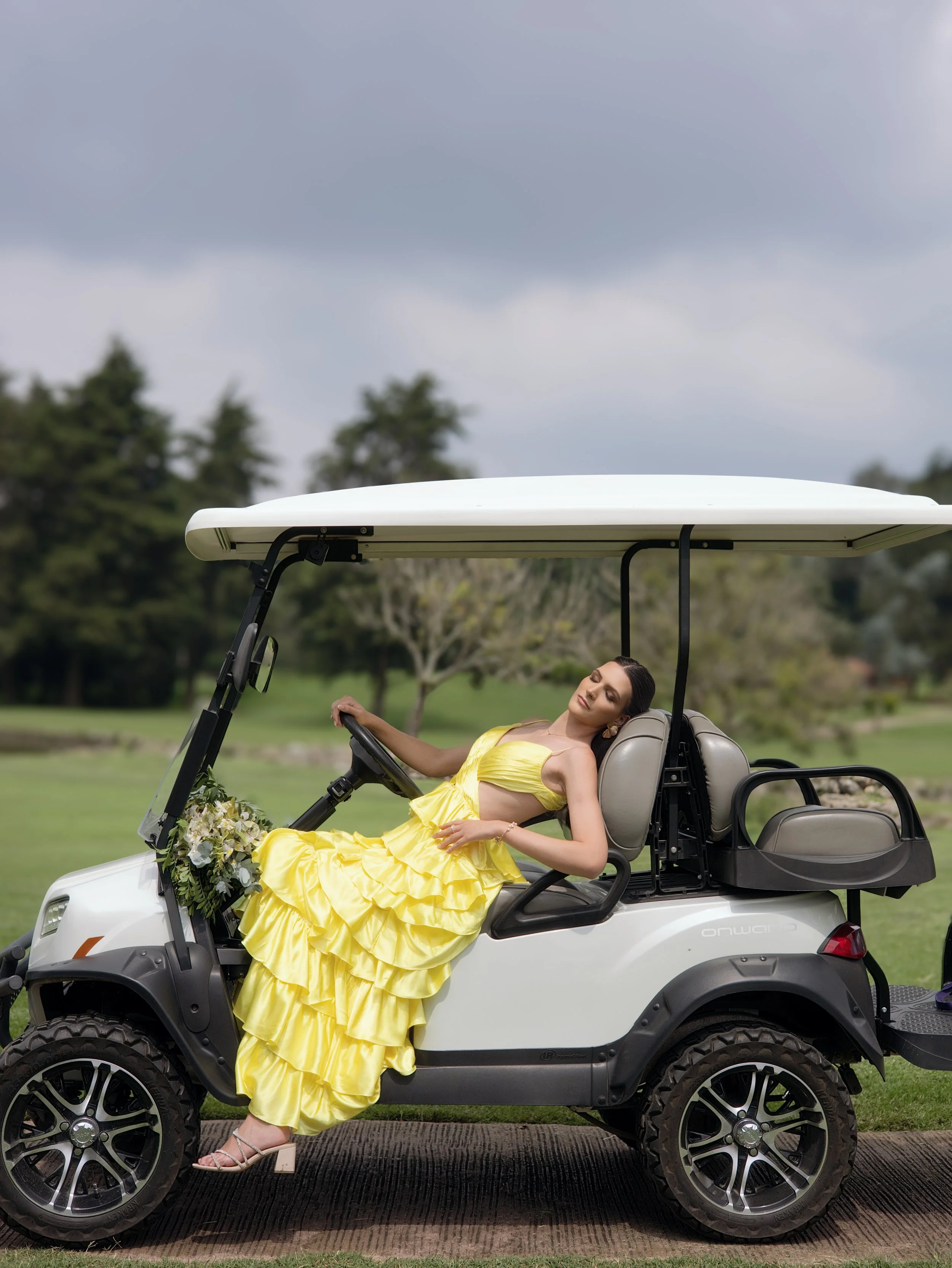 Mujer con vestido amarillo descansando en un carrito de golf decorado con flores, en un campo con césped y árboles, célebre día nublado.