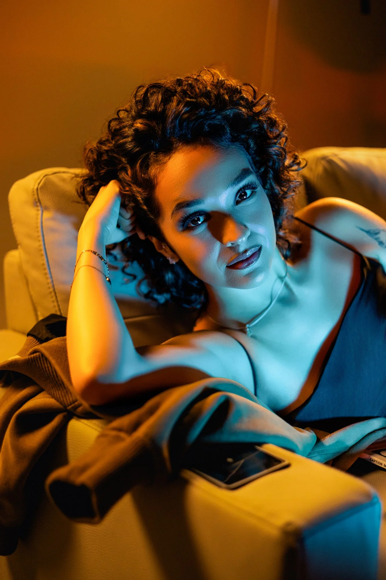 Mujer joven con cabello rizado en un sillón, mirando a la cámara, ropa oscura, en un ambiente con tonos cálidos y neón azul