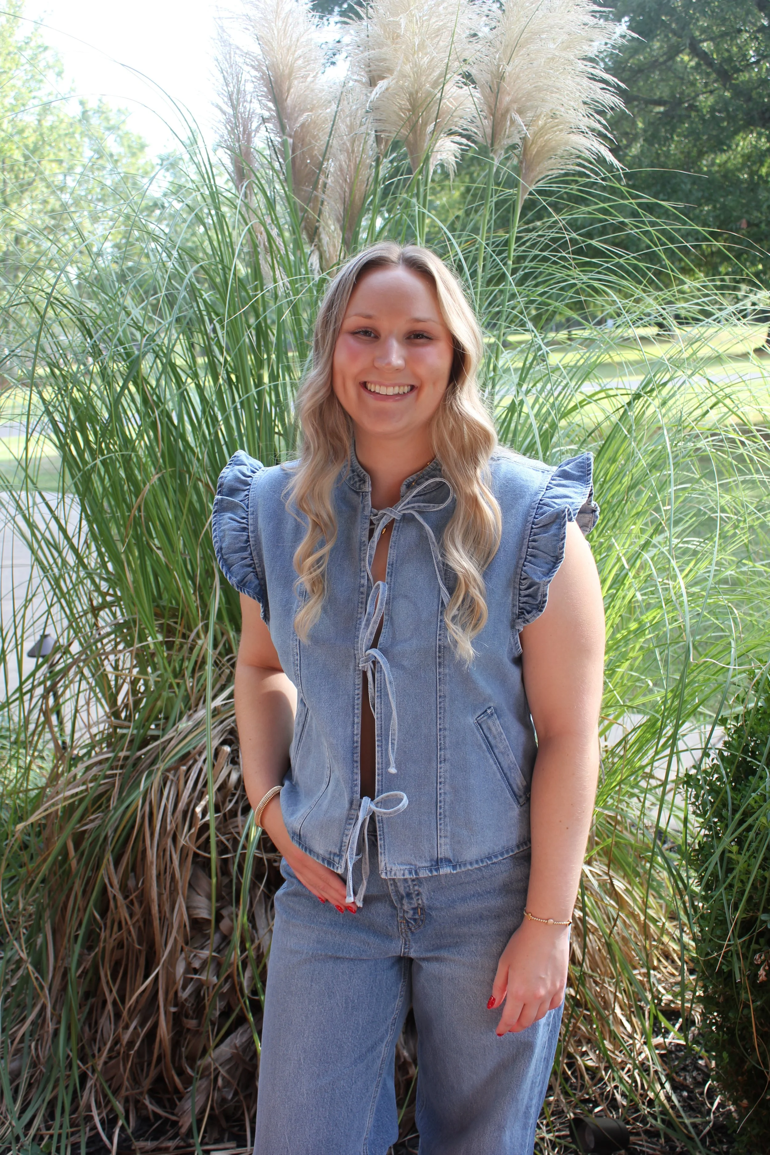 A young woman with long blonde wavy hair, smiling, stands in front of tall ornamental grass and trees on a sunny day, wearing a blue denim vest and jeans.