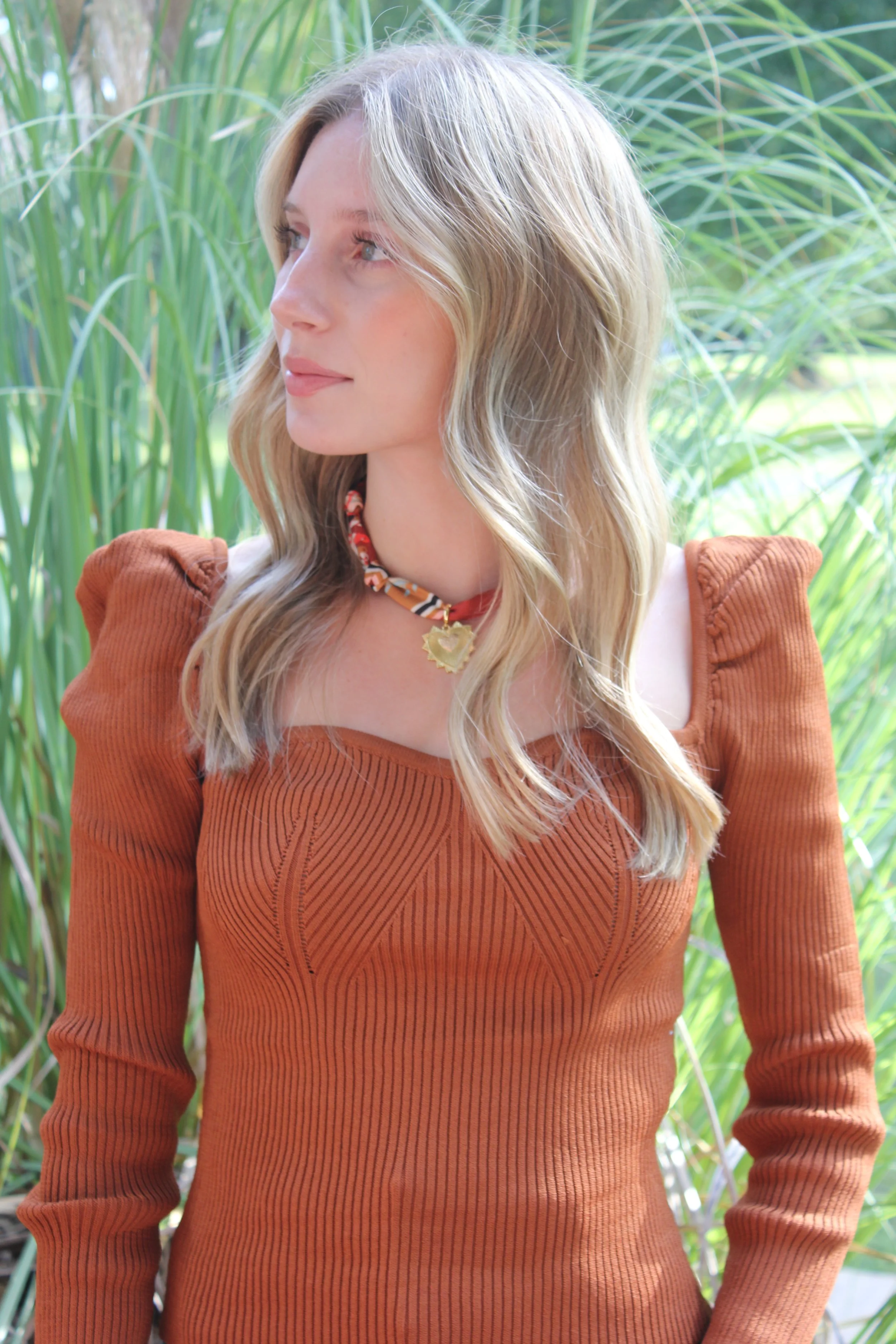 A woman with wavy blonde hair wearing a rust-colored, ribbed long-sleeve top with puffed shoulders and a patterned choker necklace with a gold pendant, standing outdoors among tall green grass.