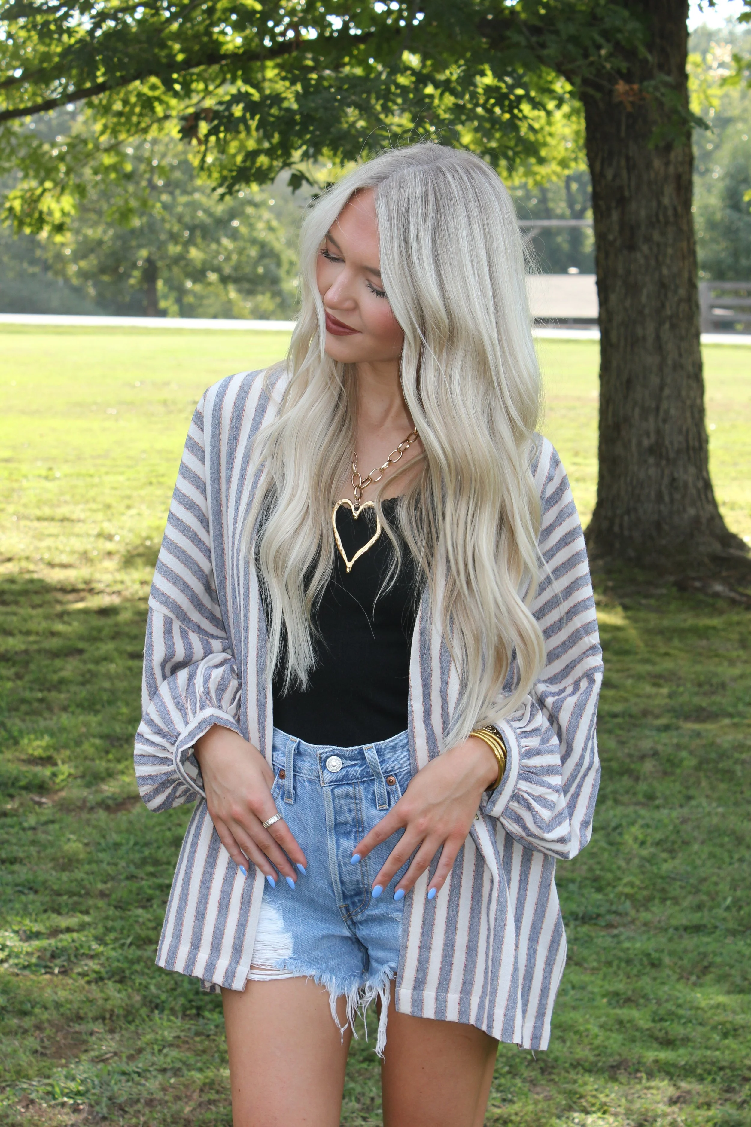 A woman with long blonde hair wearing a black top, denim shorts, and a striped blazer, standing outdoors in a park with green grass and trees.