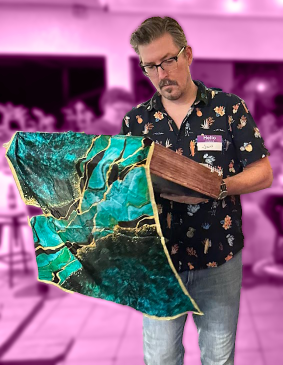 A man wearing glasses and a floral shirt holding a Blood On The Clocktower Grimoire, patterned fabric or artwork in an indoor event setting with a pink background.