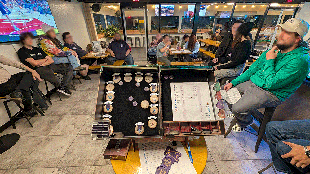 People gathered around a Blood on the Clocktower Grimoire in a bar playing a tabletop board game with various tokens and cards, while others sit on chairs and benches watching.