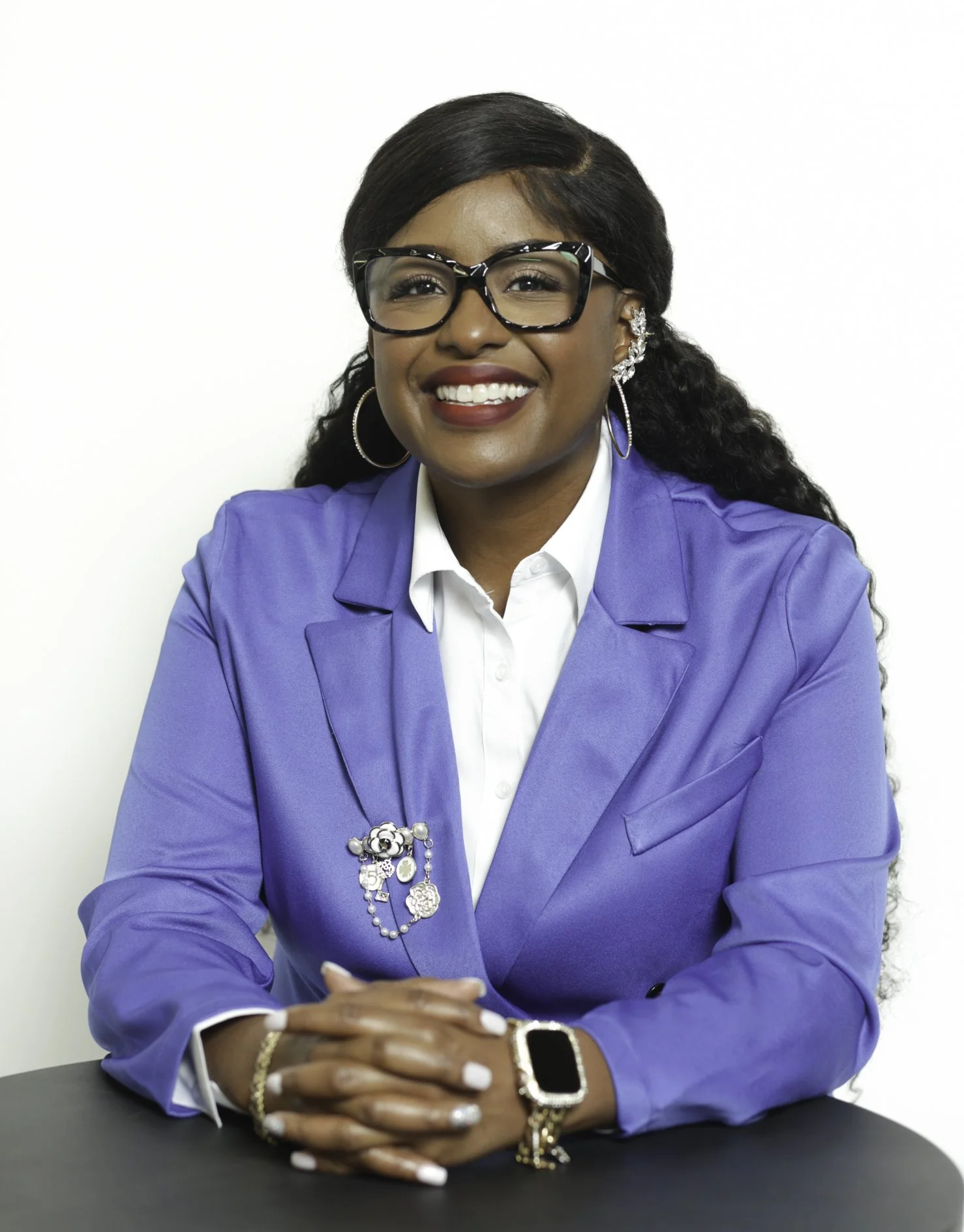 A woman sitting at a table, smiling, wearing glasses, a blue blazer with brooches, white shirt, and jewelry, against a white background.