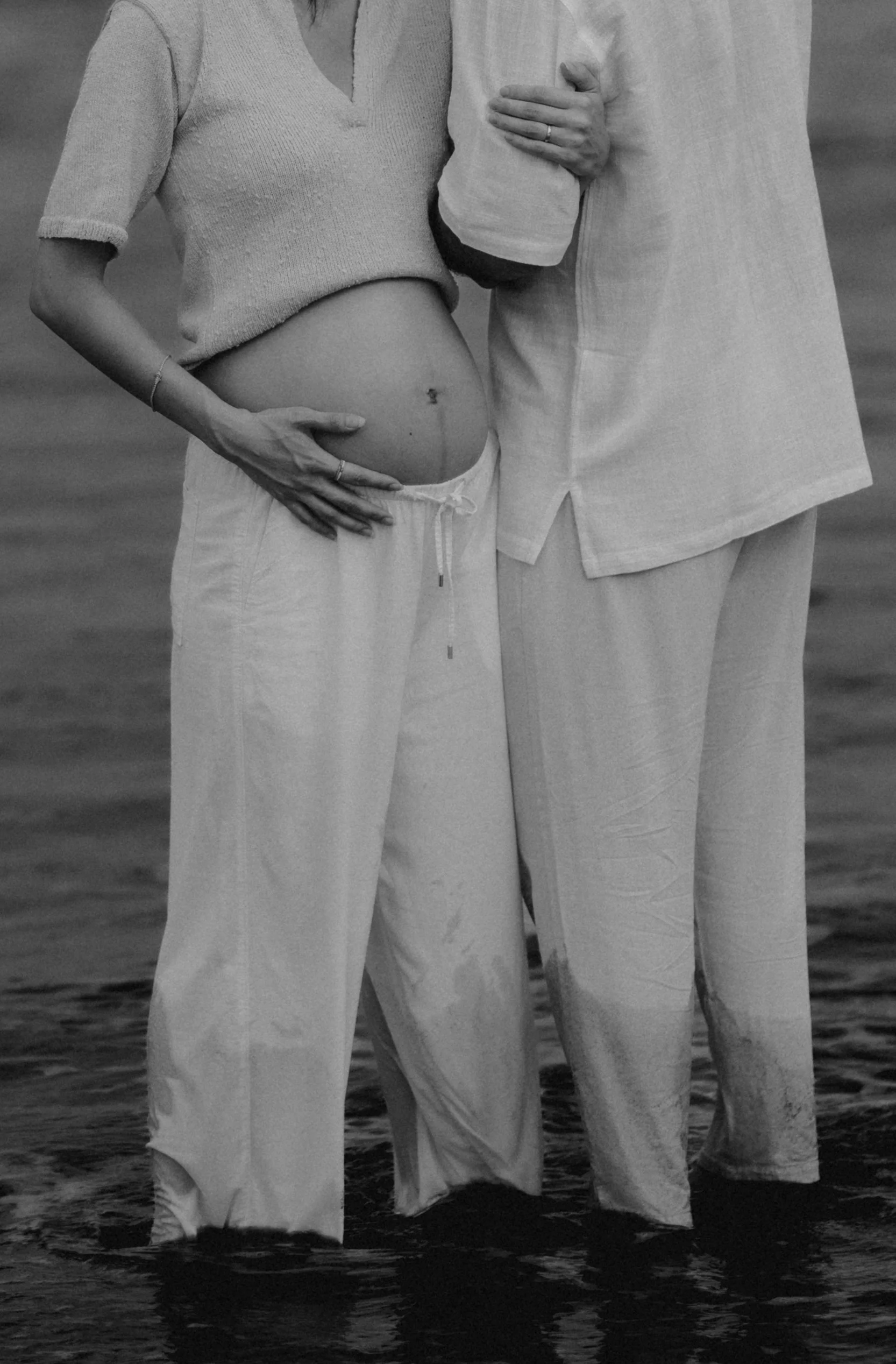 A close-up of a couple standing in water, with the woman's hand on her pregnant belly and the man's arm around her shoulder, both wearing light-colored clothing.