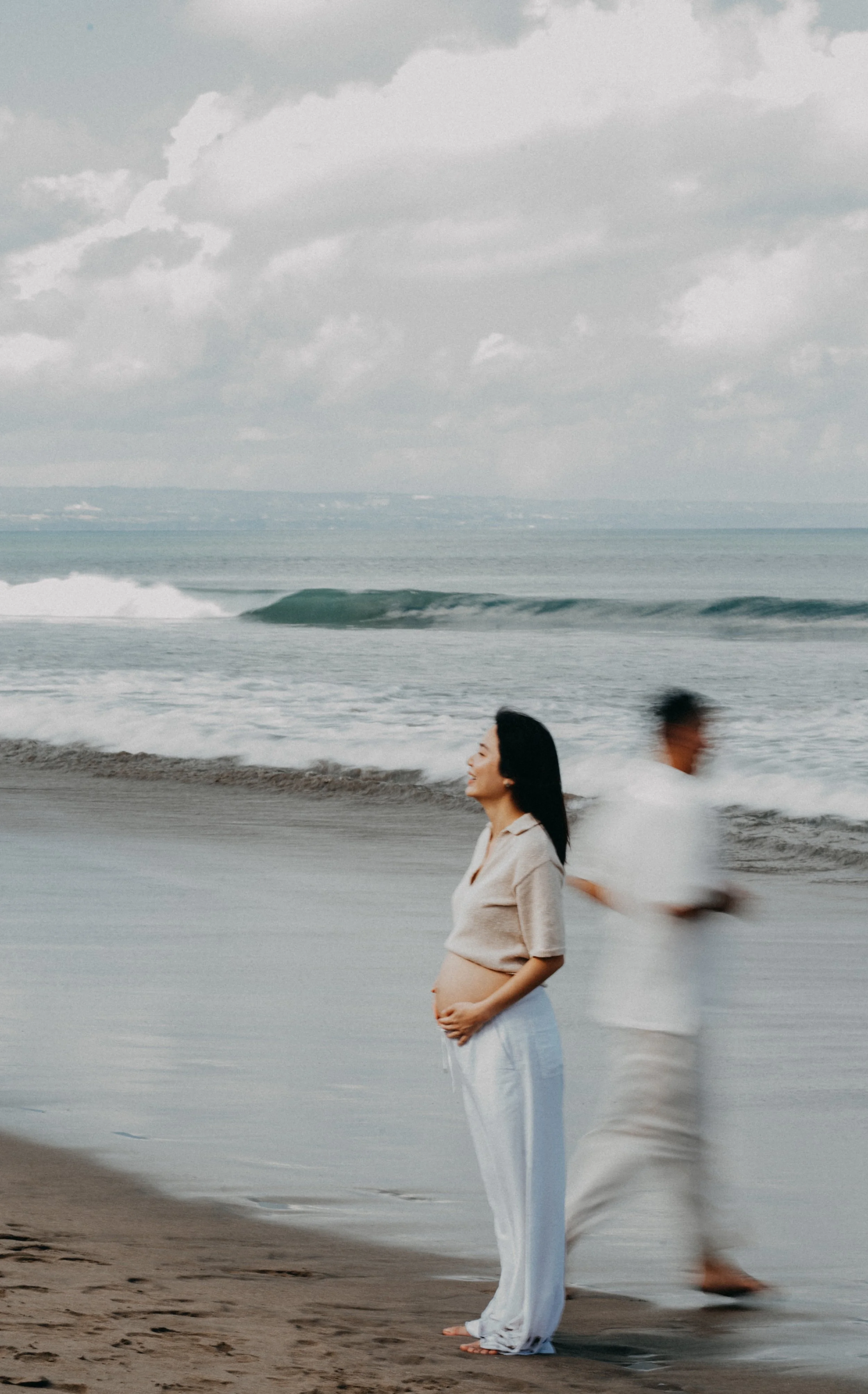A pregnant woman stands on the beach with the ocean in the background, smiling with eyes closed, while a man walks behind her. The sky is cloudy, and the woman is wearing a beige top and white pants.