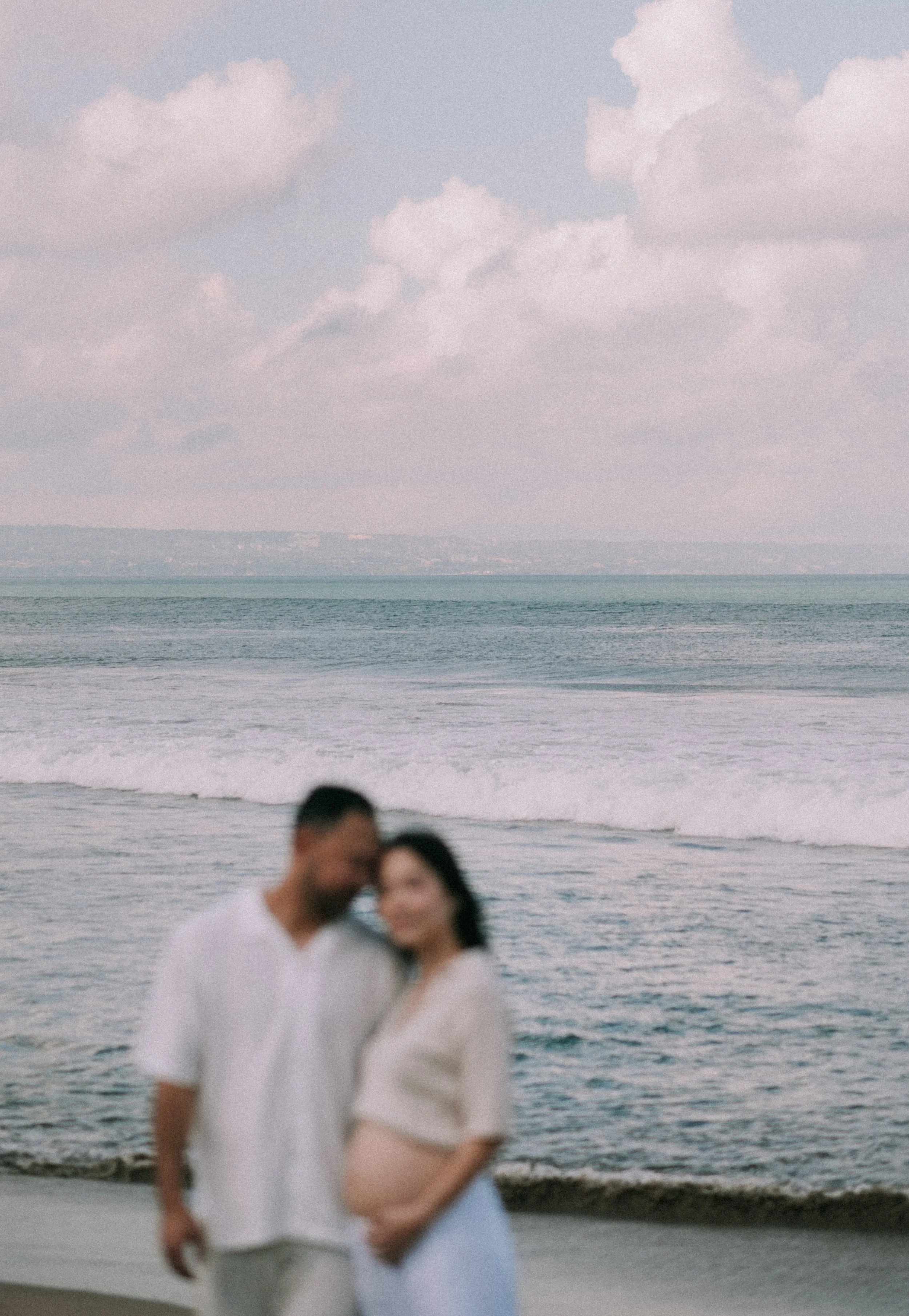 A couple stands on a beach with ocean waves and cloudy sky in the background, but their faces are blurry.