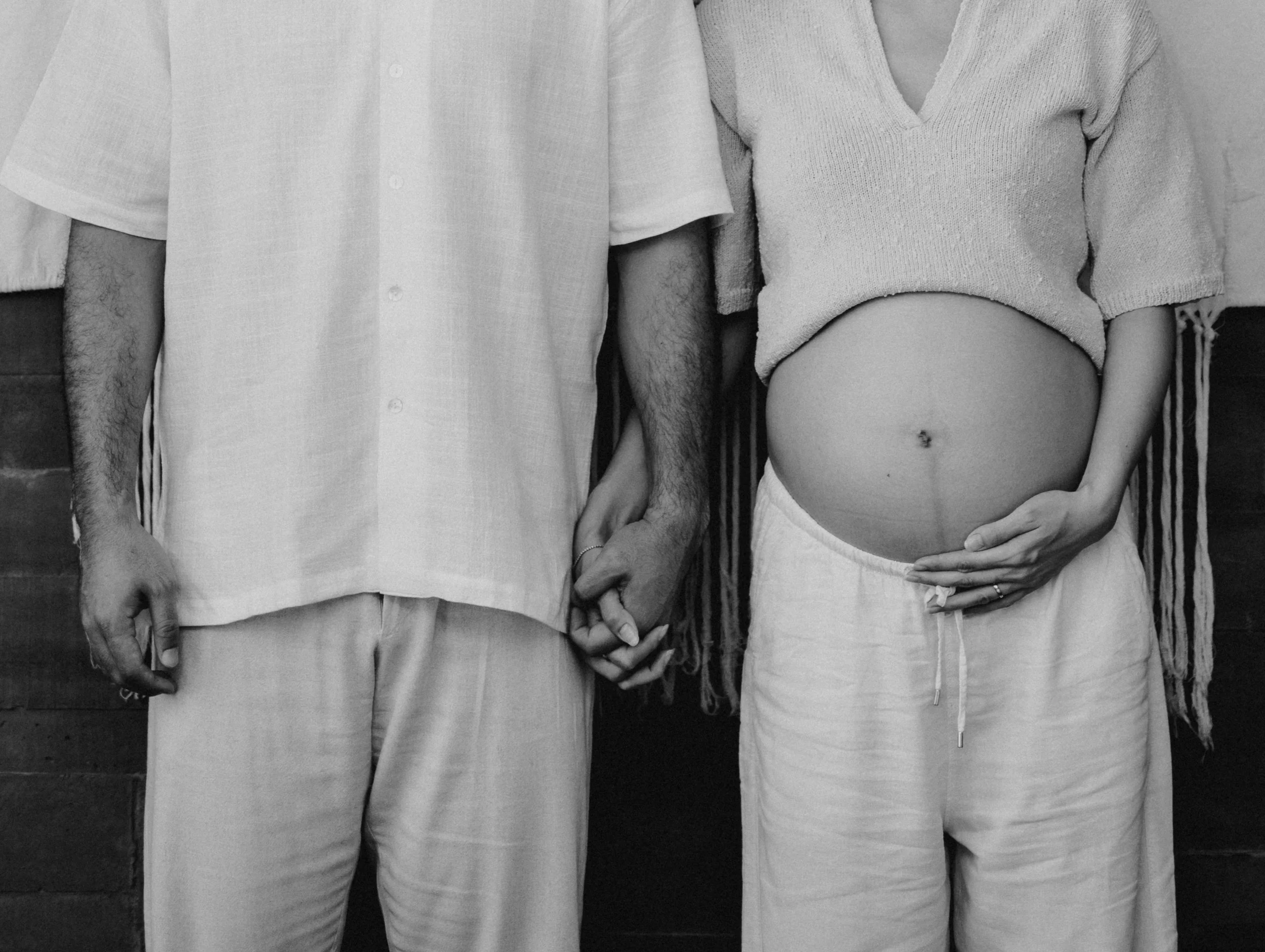 Close-up of a couple holding hands, with the woman's pregnant belly visible beneath her loose sweater, dressed in casual clothing.