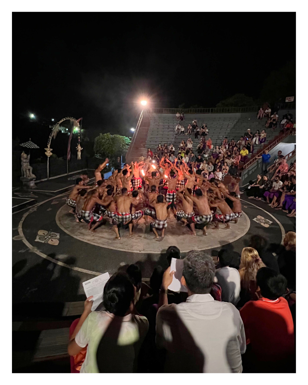 Traditional cultural performance with dancers in checkered sarongs performing a fire dance in a circular formation at night, surrounded by an audience seated in an open-air amphitheater.
