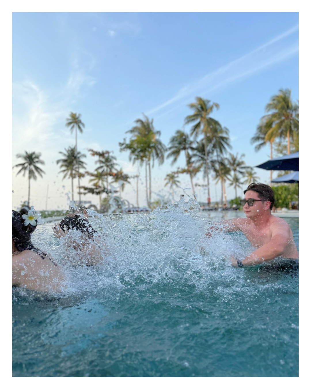 People playing in a swimming pool with palm trees and a clear blue sky in the background.
