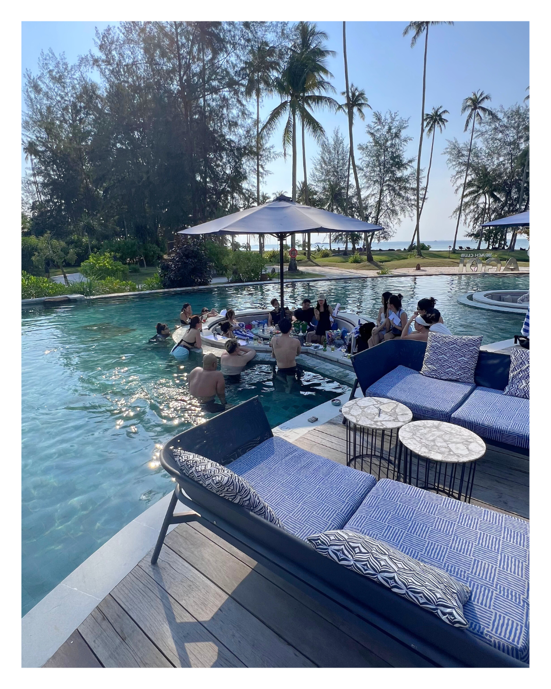 People gathered in and around a swimming pool with loungers, umbrellas, and lush tropical trees in the background.