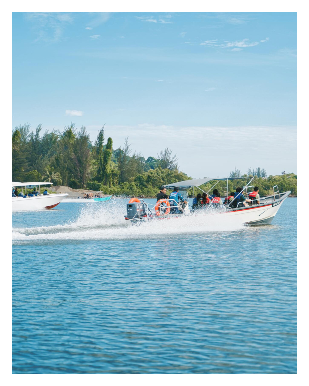 People riding in a motorboat on a calm lake with trees and blue sky in the background.