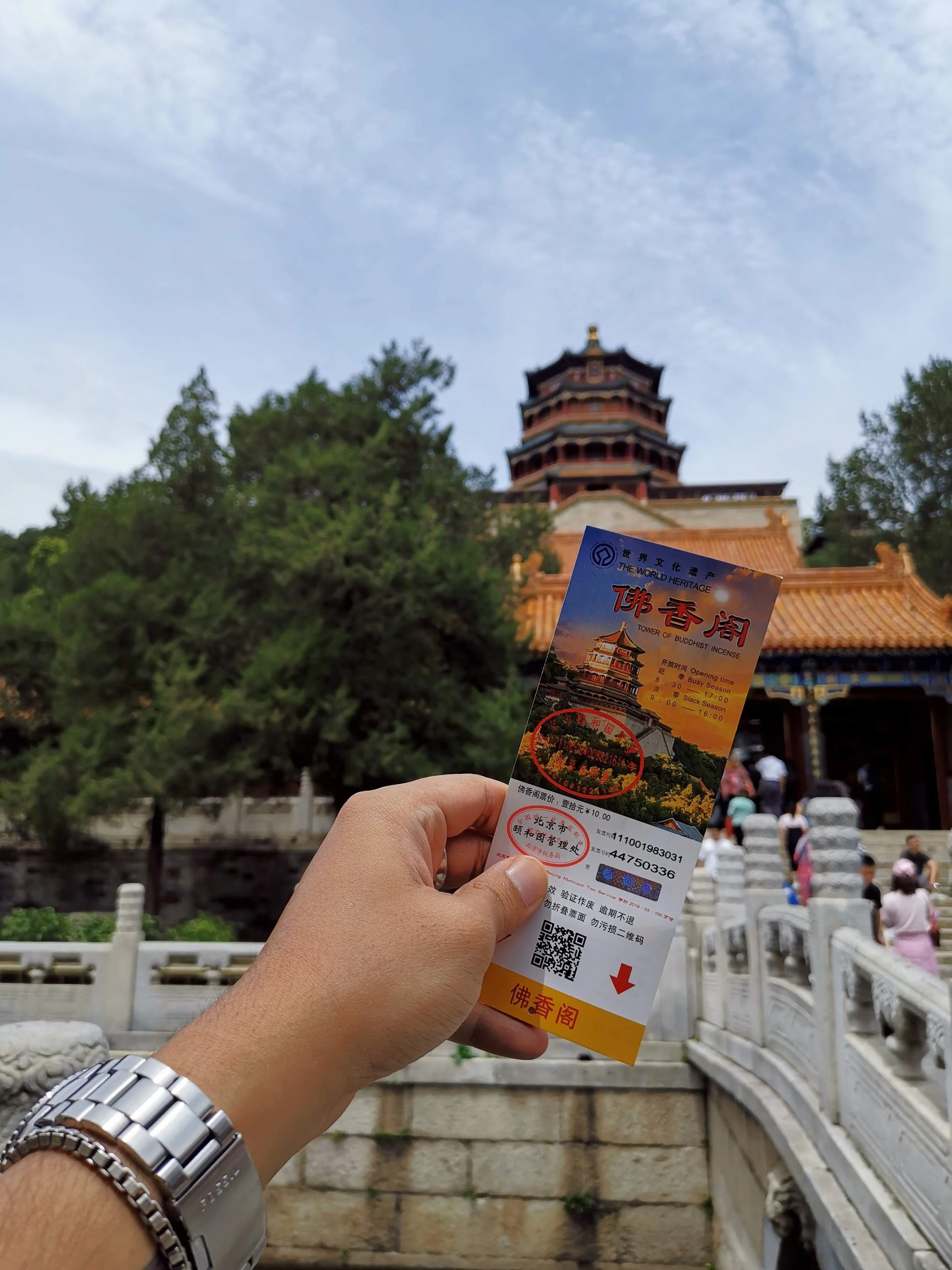 A person holding a ticket for the Tower of Buddhist Incense in front of a traditional Chinese pagoda with people climbing stairs near it.