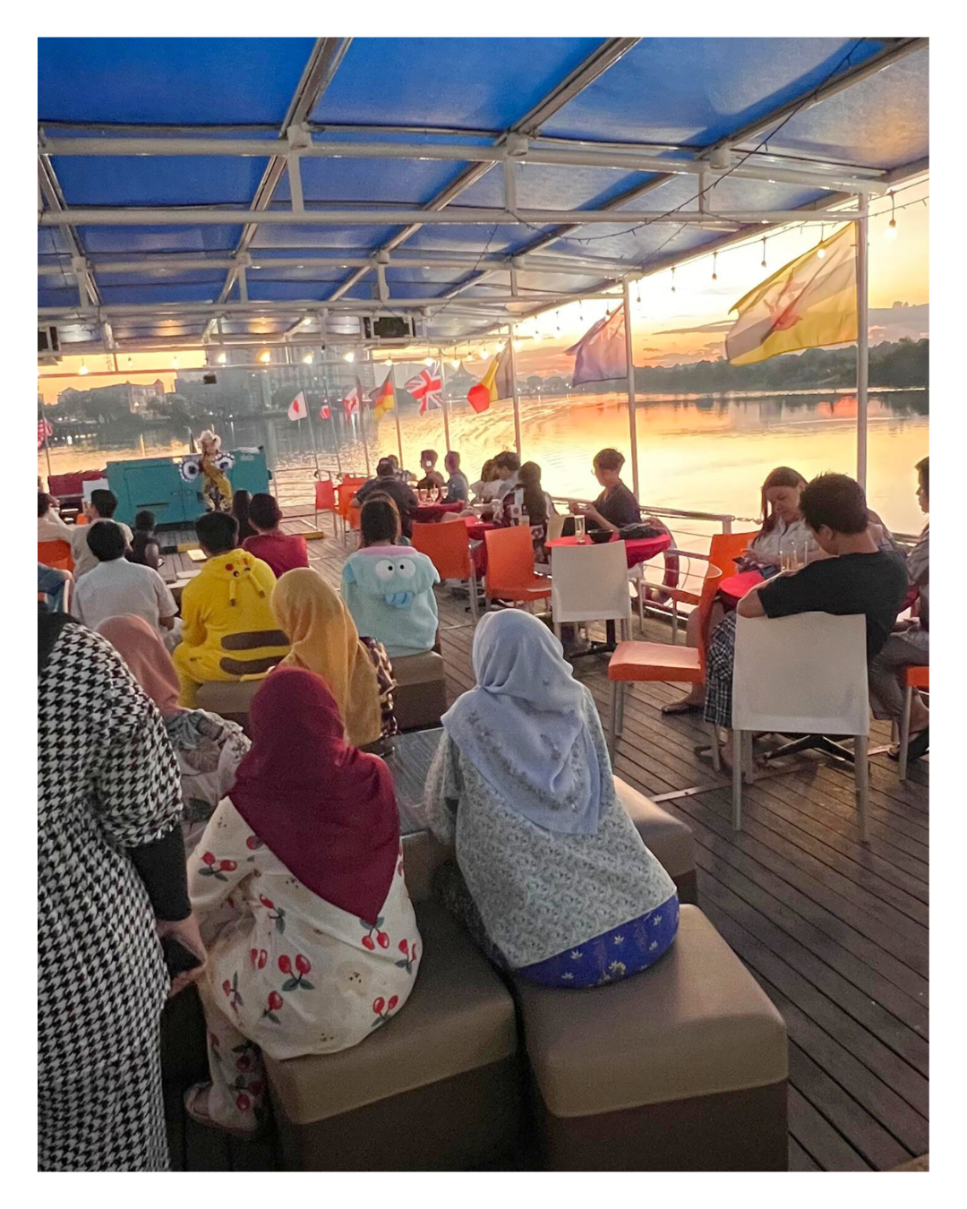 People dining on a boat at sunset, overlooking a river with flags from different countries, with some people wearing costumes.