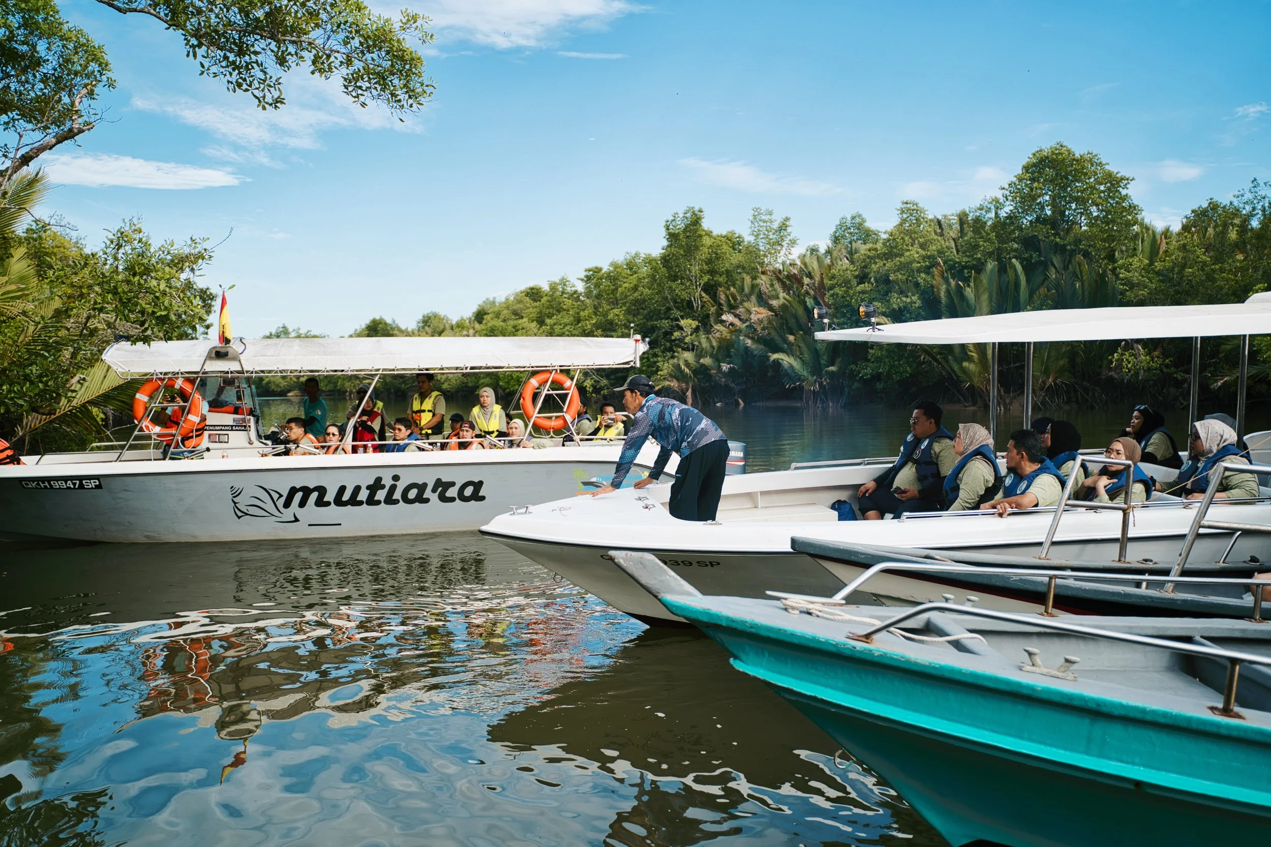 People on two boats in a river surrounded by lush green trees under a clear blue sky.