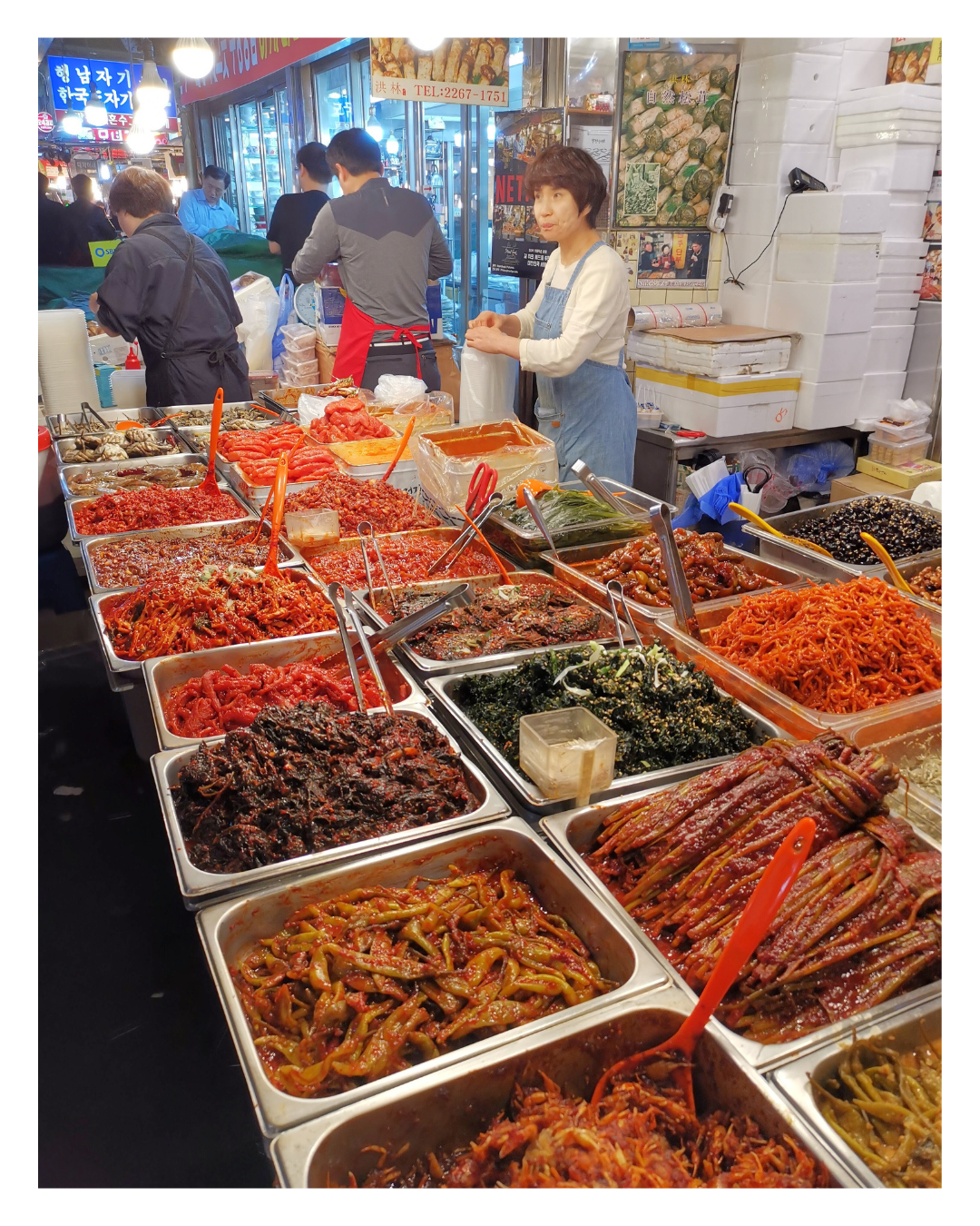 Food stall selling various types of Korean side dishes, including kimchi, seasoned vegetables, and seafood, with vendors preparing and serving customers in a busy market.