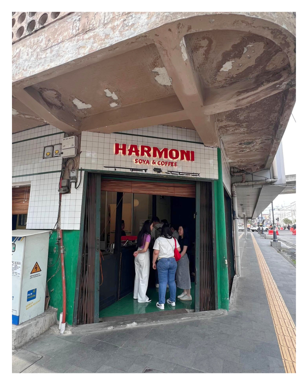A small cafe called Harmoni Soya & Coffee with people gathered inside, located on a busy street corner with a sidewalk and traffic visible outside.