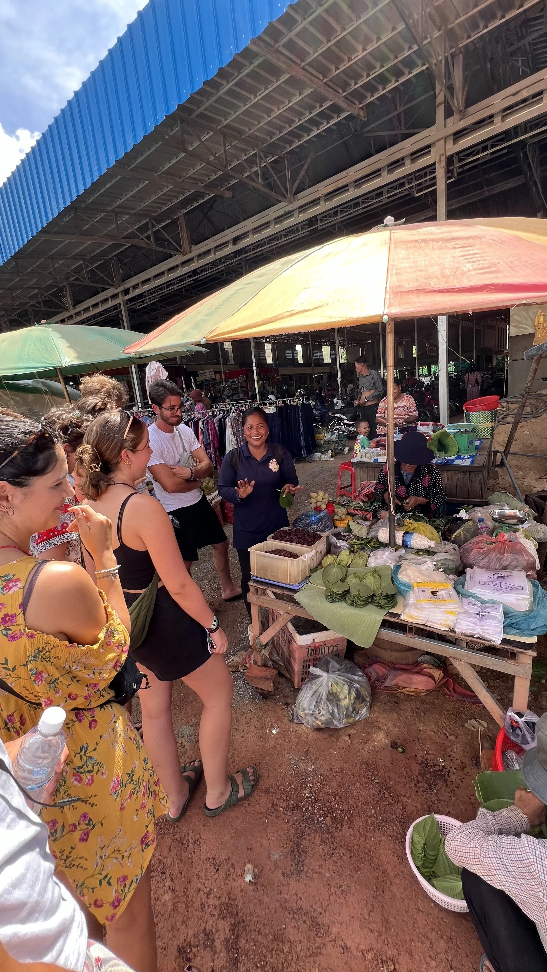 People shopping and talking at an outdoor market stall with fruits, vegetables, and other goods under colorful umbrellas near a covered market building.