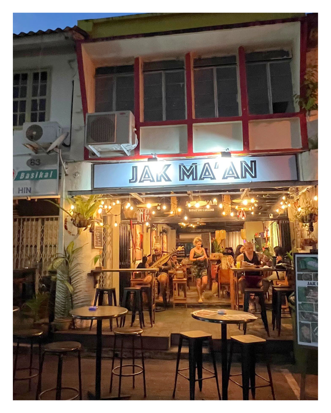 A nighttime scene of a lively restaurant or bar with outdoor seating, string lights, and people dining inside. The sign above the entrance reads 'JAK MA'AN'.