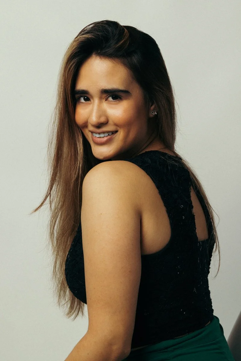 A young woman with long brown hair, smiling and looking over her shoulder, wearing a black sleeveless top.
