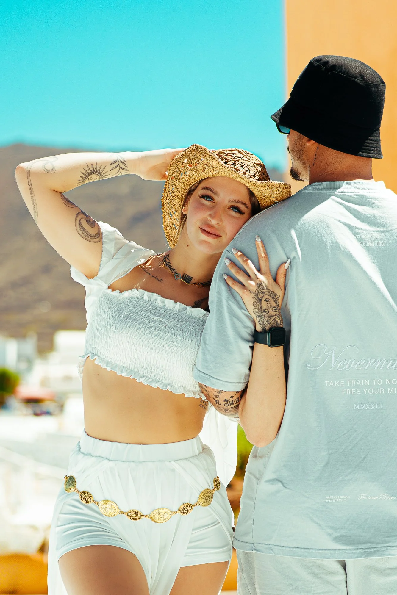 A young woman wearing a straw hat and white crop top, posing with a man wearing a bucket hat, against a desert landscape with mountains and colorful sky.