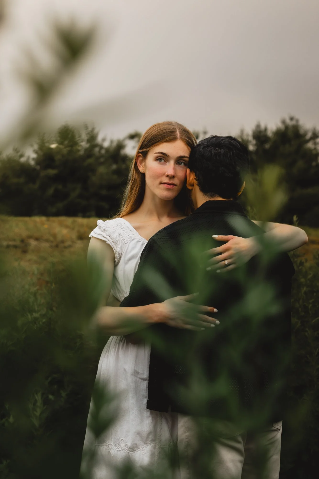 A young woman with auburn hair and a white dress embraces a man with dark hair and a black shirt outdoors, with greenery and trees in the background.