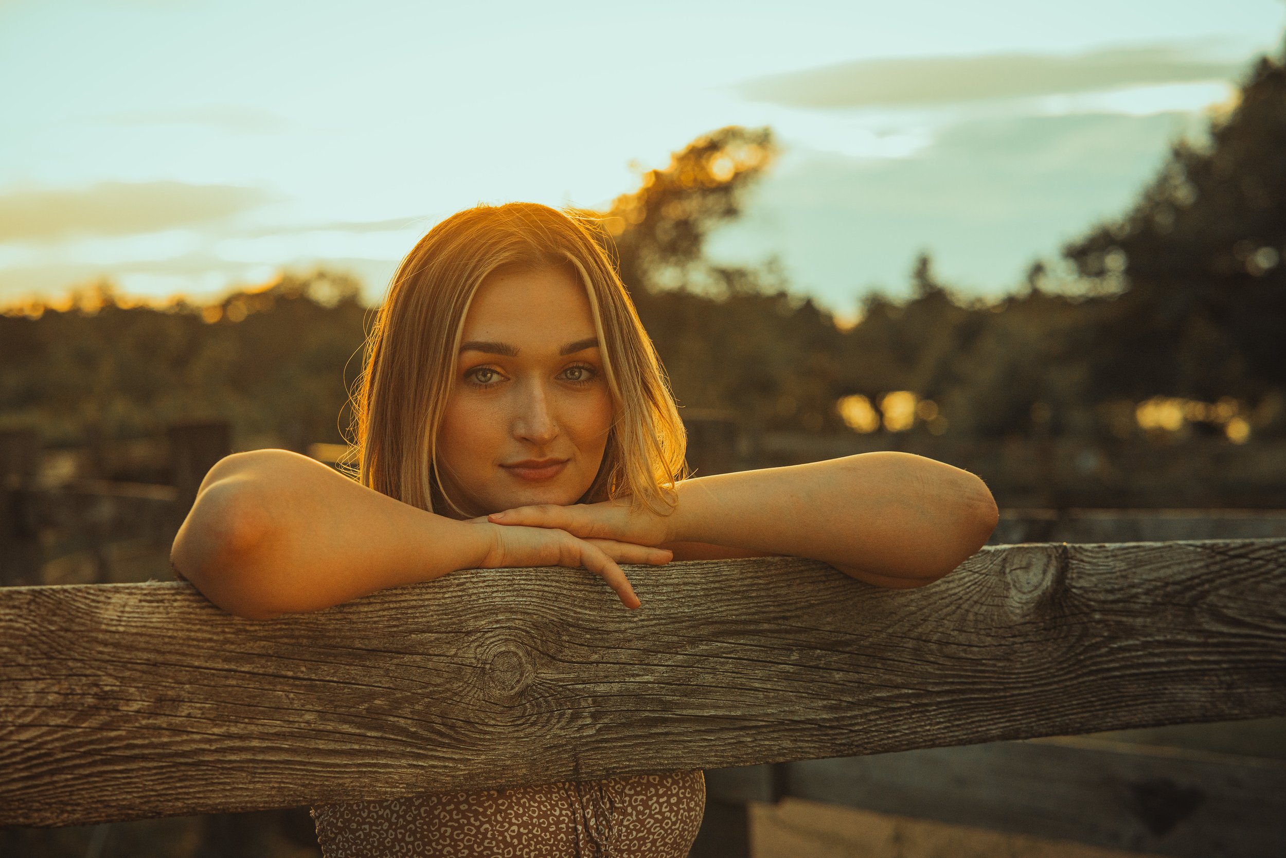 A woman with light brown hair leans on a wooden fence during sunset, with a blurred background of trees and sky.