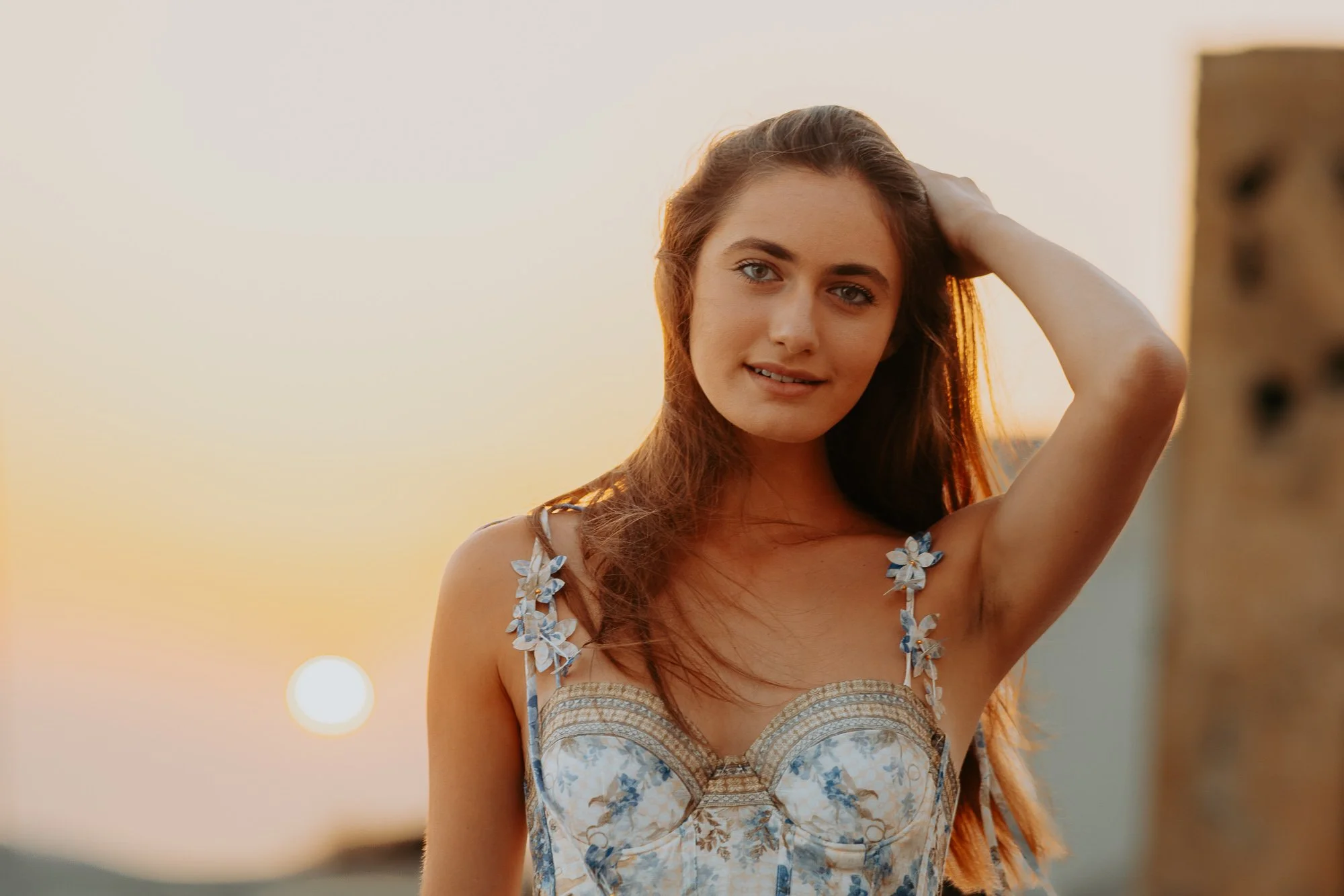 Nicole Elizabeth Berger with long brown hair wearing a floral dress, standing outdoors at sunset, smiling gently with her hand behind her head.