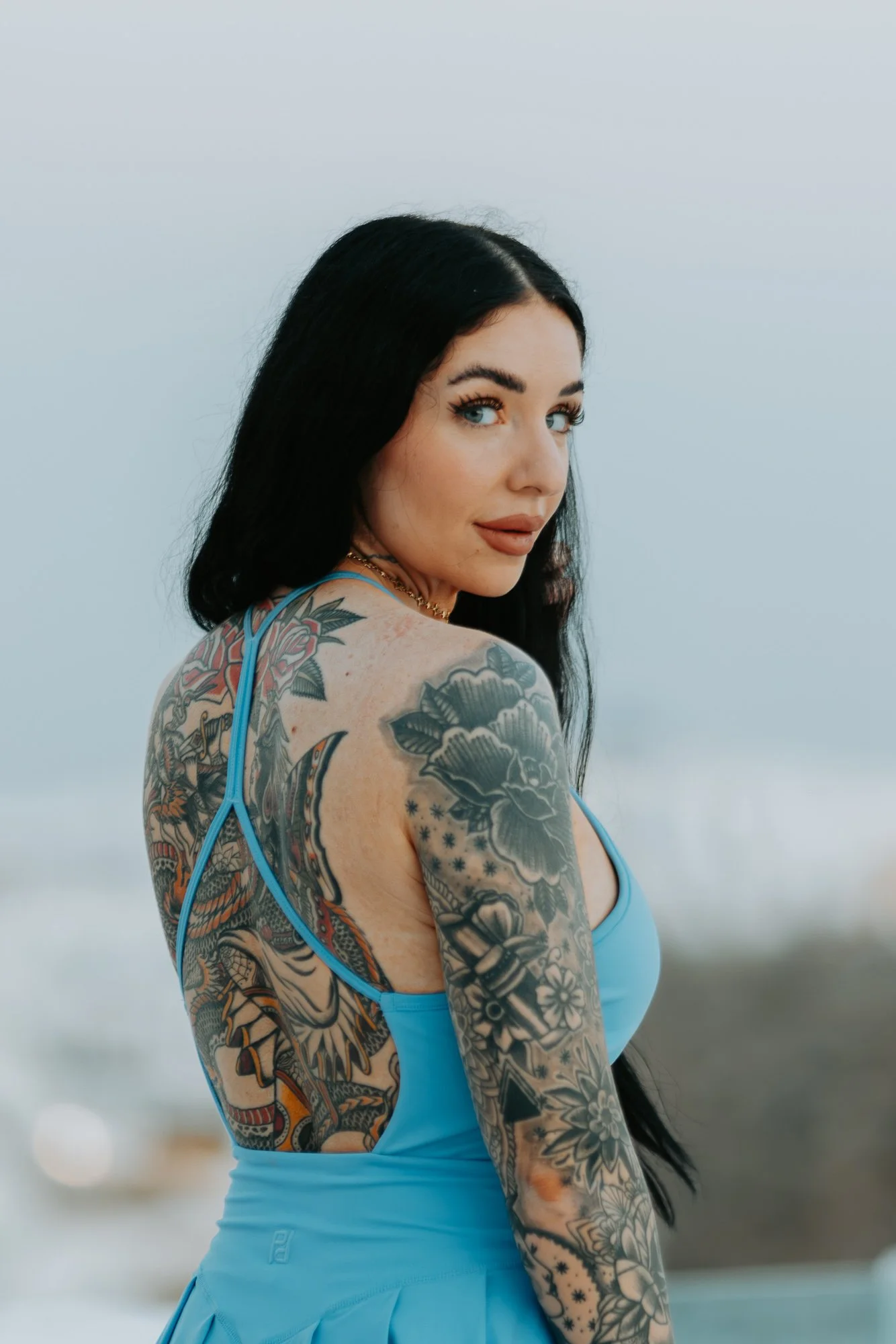 A woman with long dark hair and tattoos on her arm and shoulder, wearing a blue dress, standing outdoors with a blurred ocean background, looking over her shoulder.