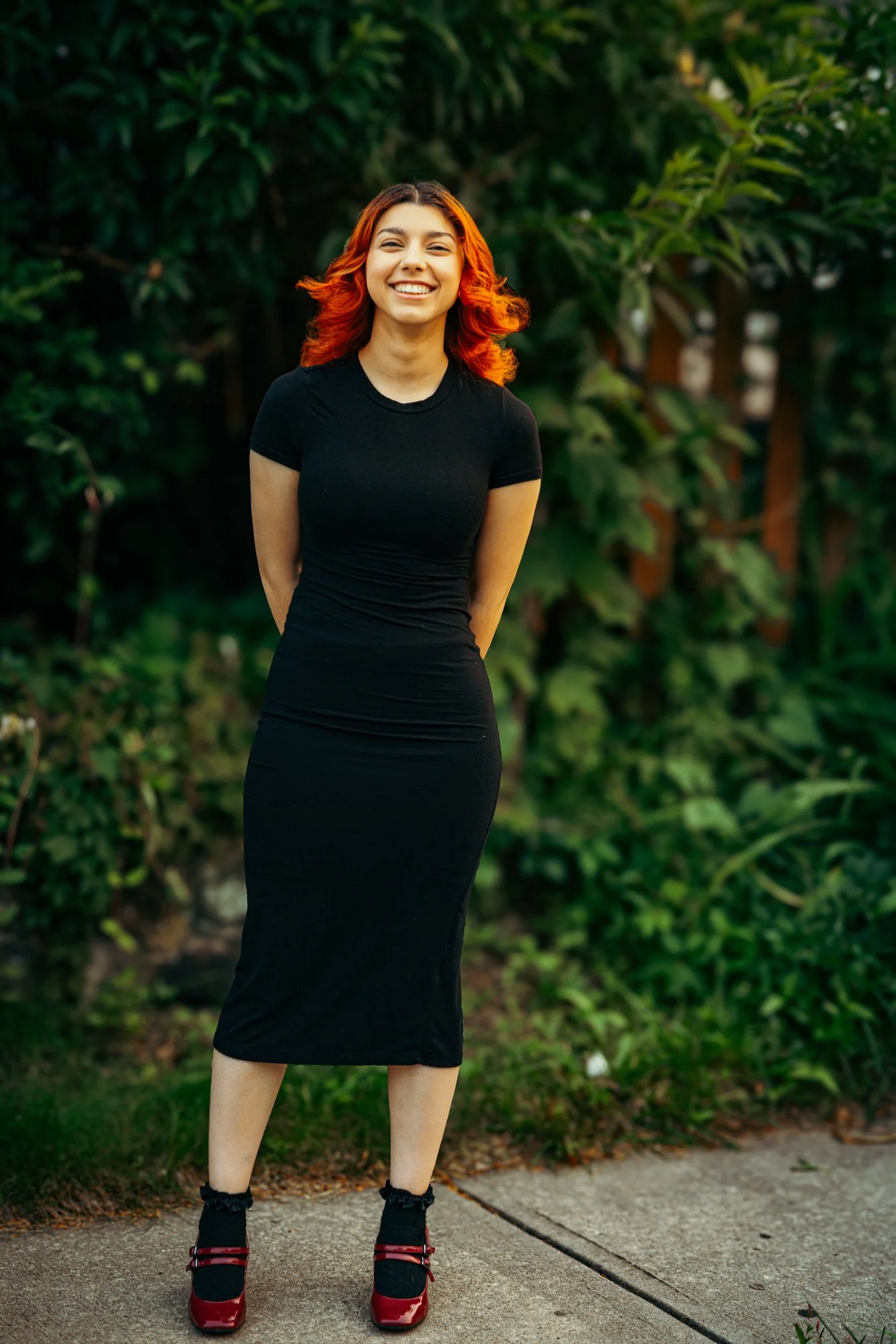 A woman with red hair standing outdoors on a sidewalk, smiling, wearing a black dress and red shoes with black socks, with green foliage in the background.
