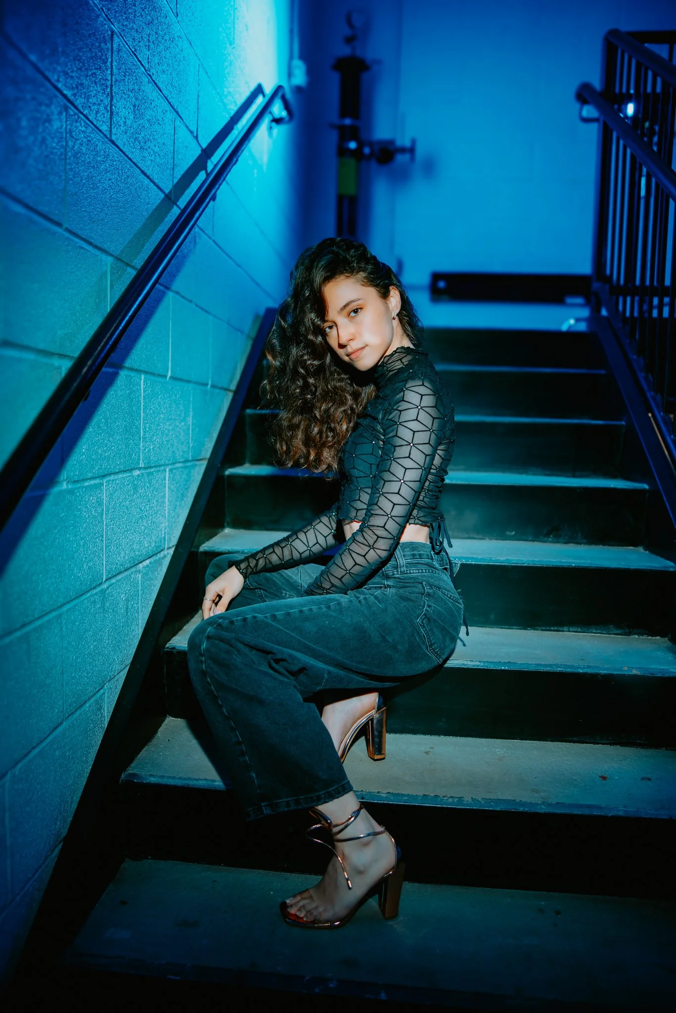 Young woman with curly hair sitting on stairs, wearing a black fishnet-style top, high heels, and dark jeans, illuminated by blue lighting.