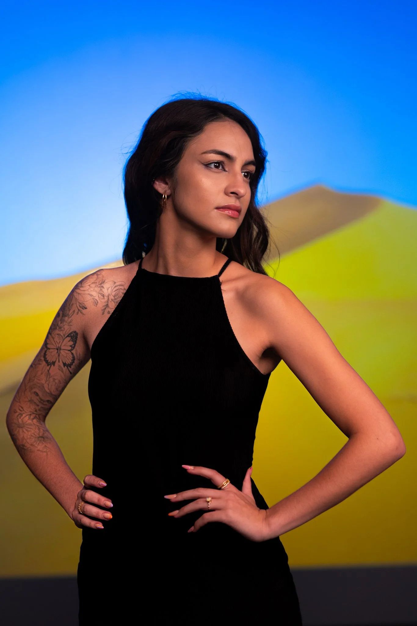 A woman with dark hair, wearing a sleeveless black dress, standing against a colorful abstract background. She has tattoos on her left arm and accessories on her fingers.