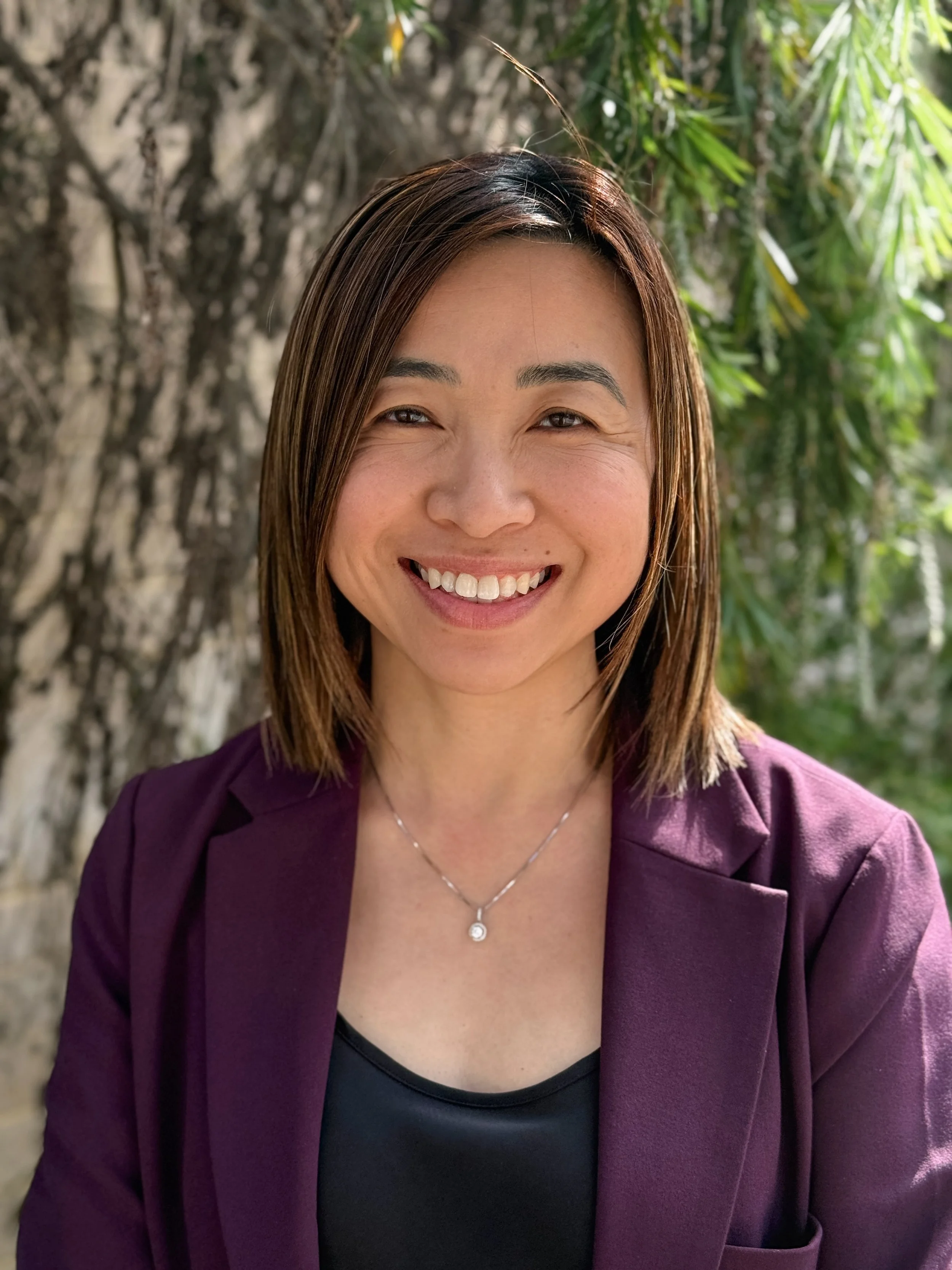 A woman with shoulder-length brown hair smiling outdoors in front of a tree with green leaves, wearing a purple blazer, a black top, and a delicate necklace.