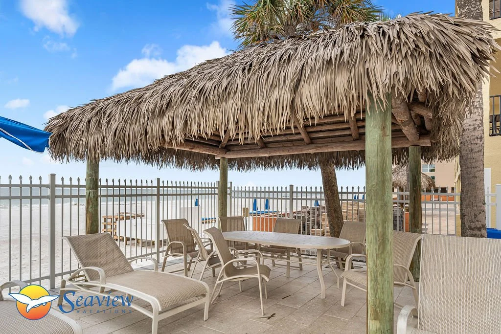 Beachside seating area with lounge chairs under a tiki hut,  ocean view in the background.