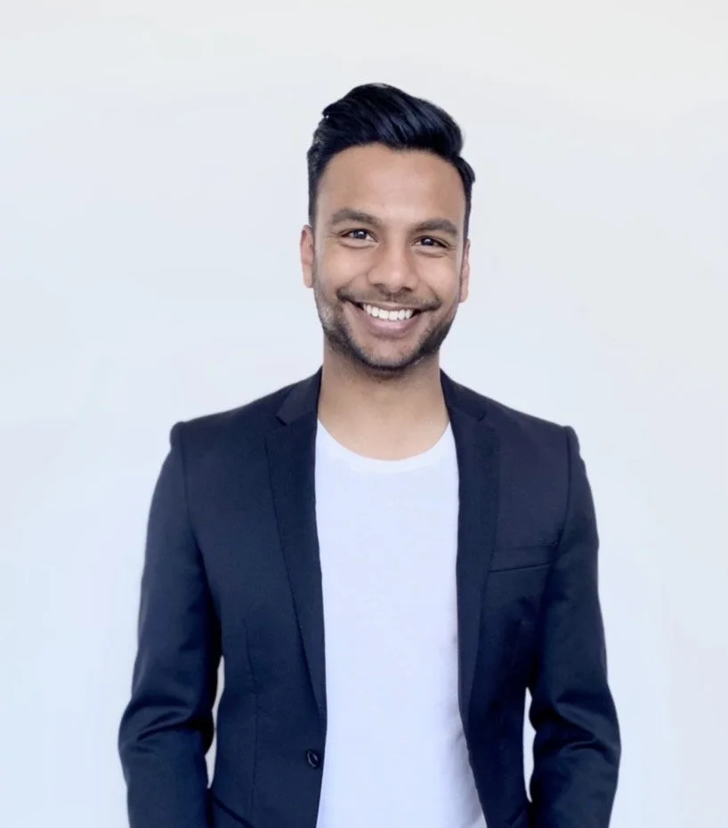 A smiling man with dark hair, wearing a dark blazer and a white shirt, standing against a plain light background.